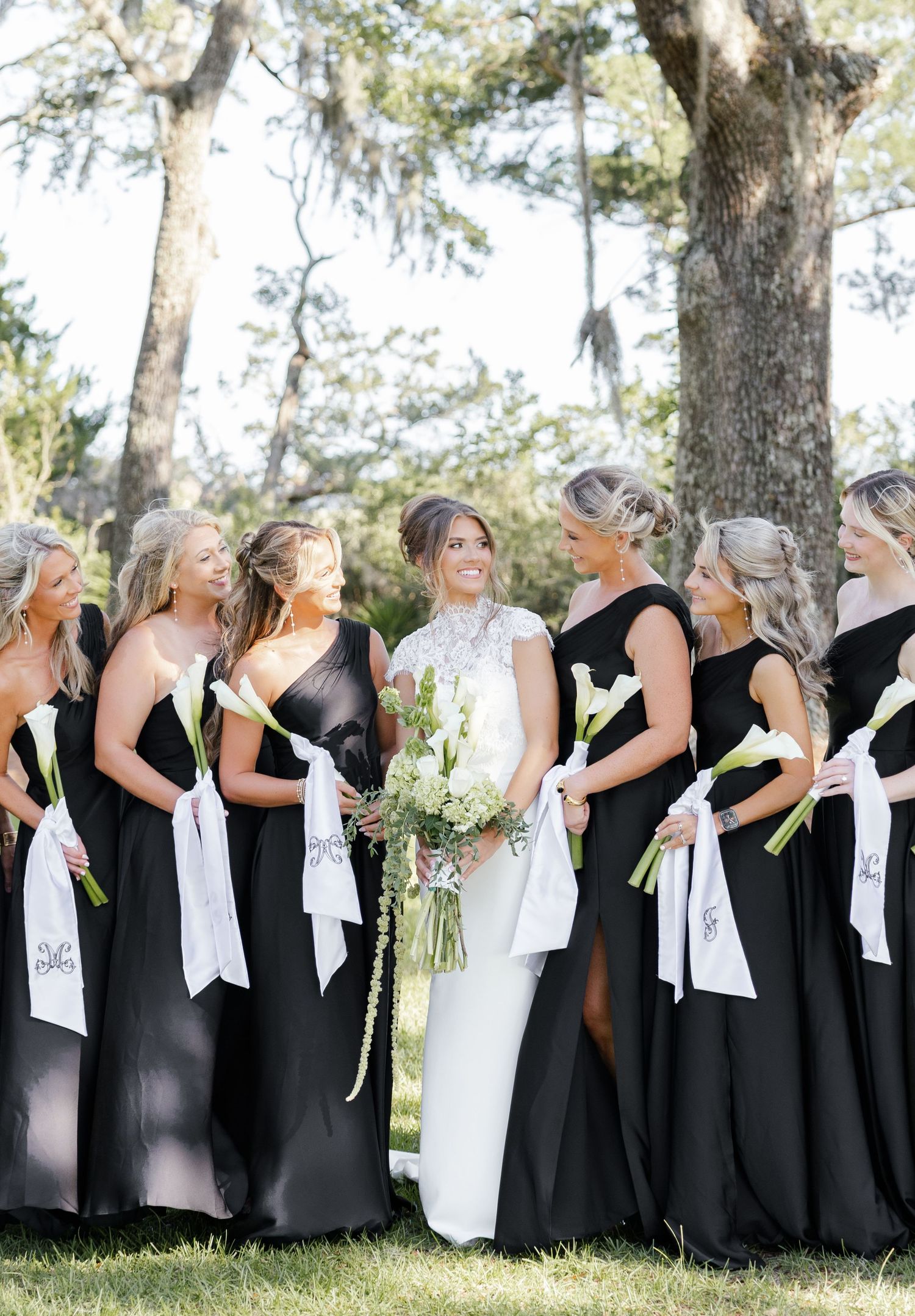 Bride in white gown with bridesmaids in black dresses holding white bouquets outdoors under trees at elegant wedding.