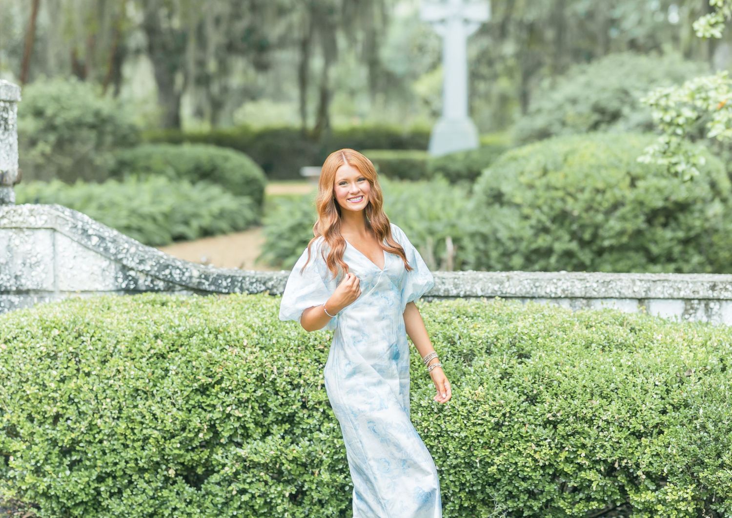 Someone in a white dress spins gracefully in front of manicured garden hedges on a sunny summer day.