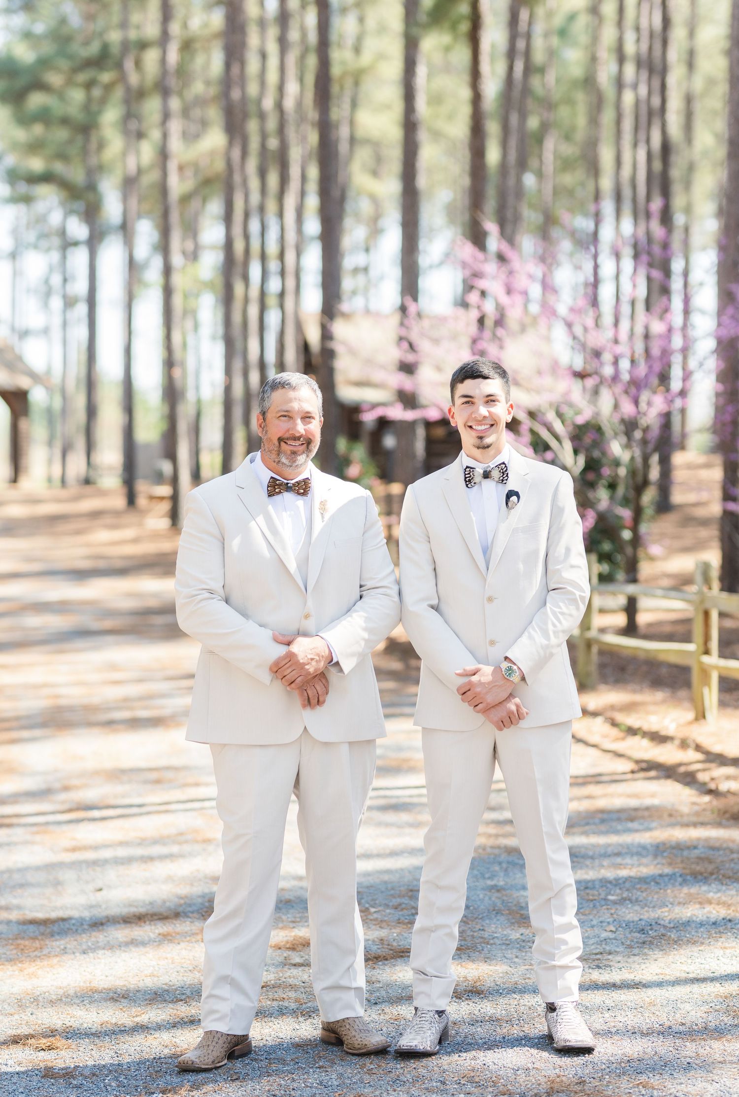 Two men in matching cream suits with bow ties smile together on gravel path at wedding venue.