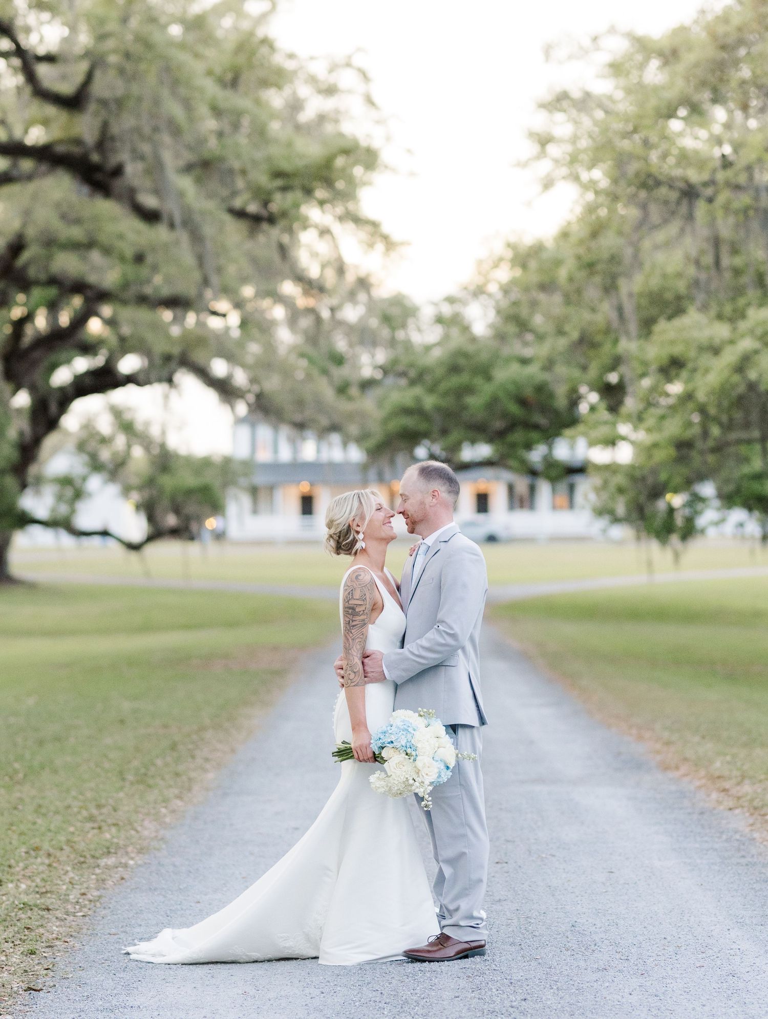 Bride and groom kissing on a scenic driveway with Spanish moss trees and white plantation house backdrop.