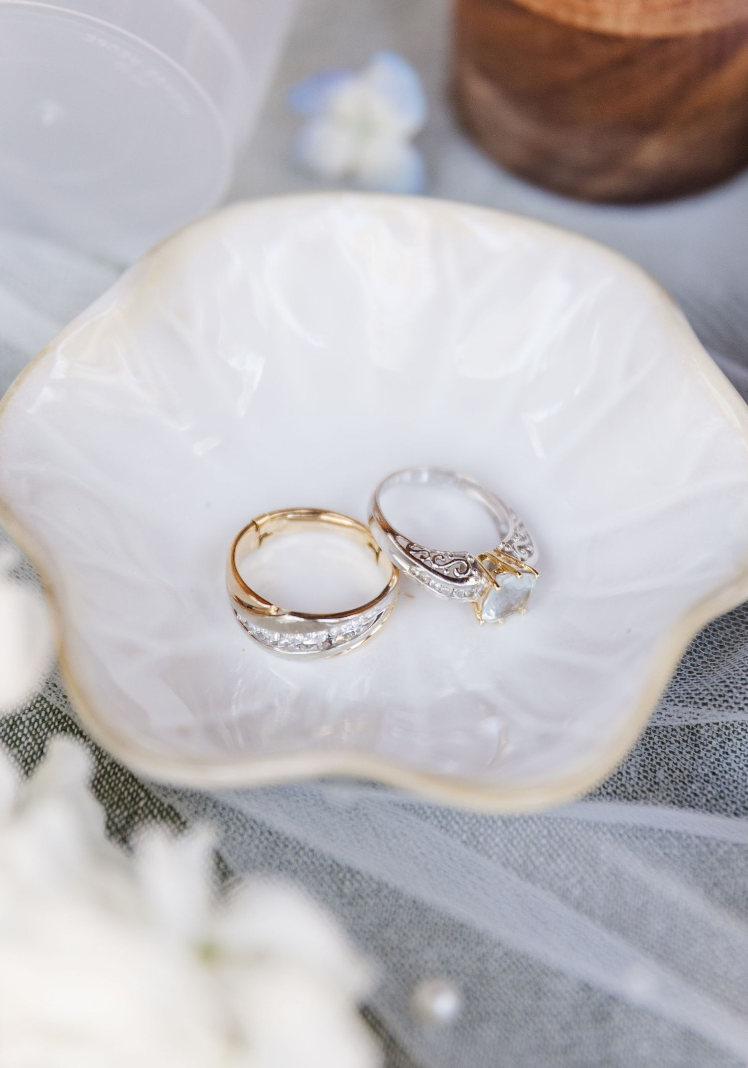 Two wedding rings rest in a white ceramic shell dish, one gold band and one diamond engagement ring.