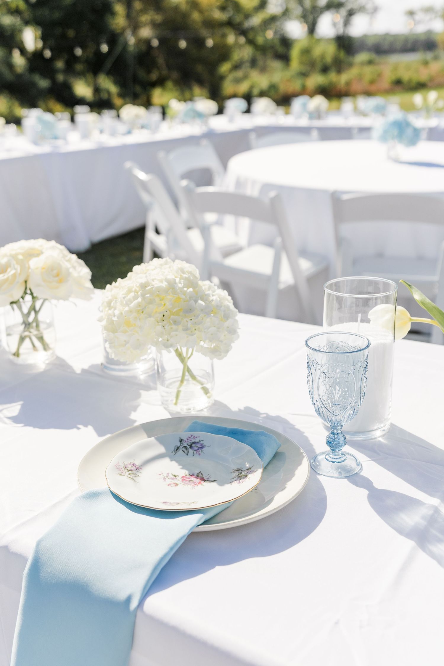 Elegant outdoor wedding reception table with white hydrangeas, blue glassware, and light blue napkins on white linen.