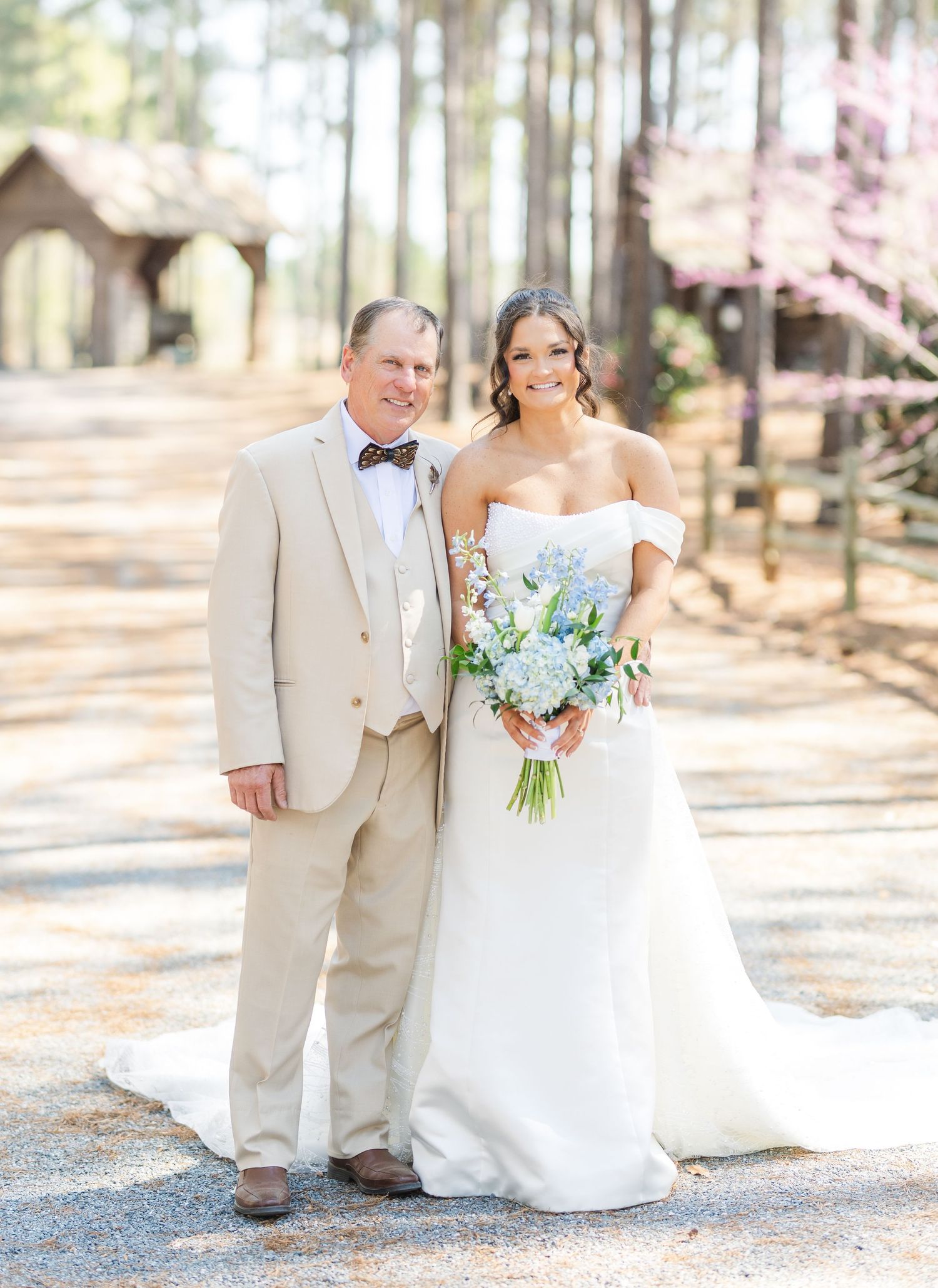 Bride in off-shoulder white gown holds blue bouquet posing with older gentleman in tan suit outdoors.