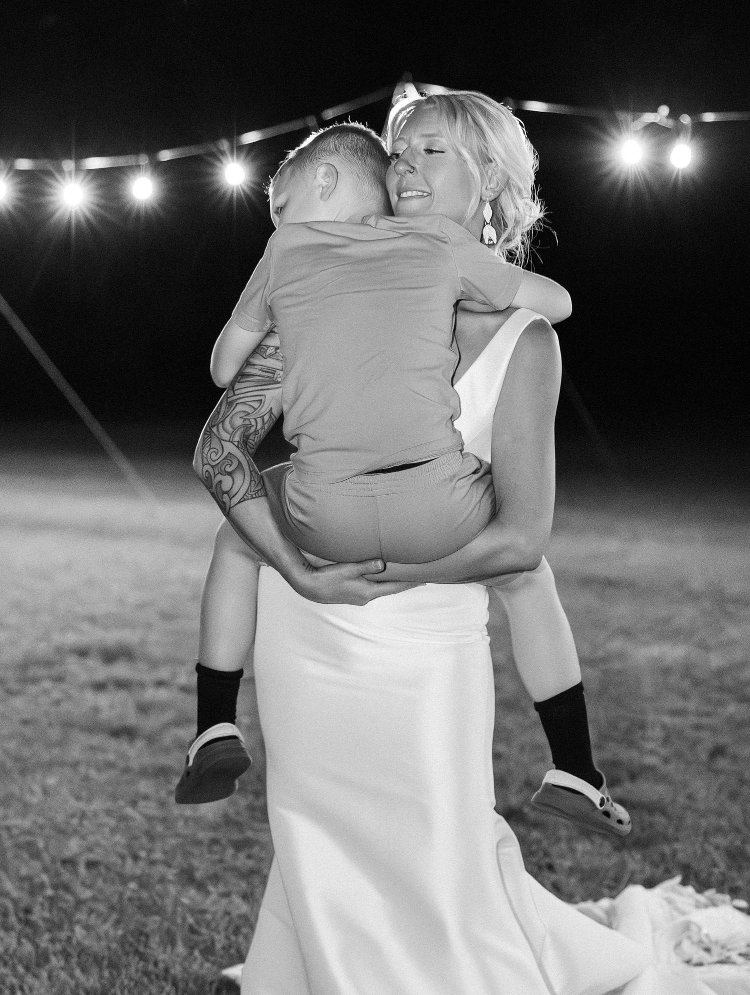 Black and white photo of a smiling bride in a white gown lifting a young child at a nighttime reception.