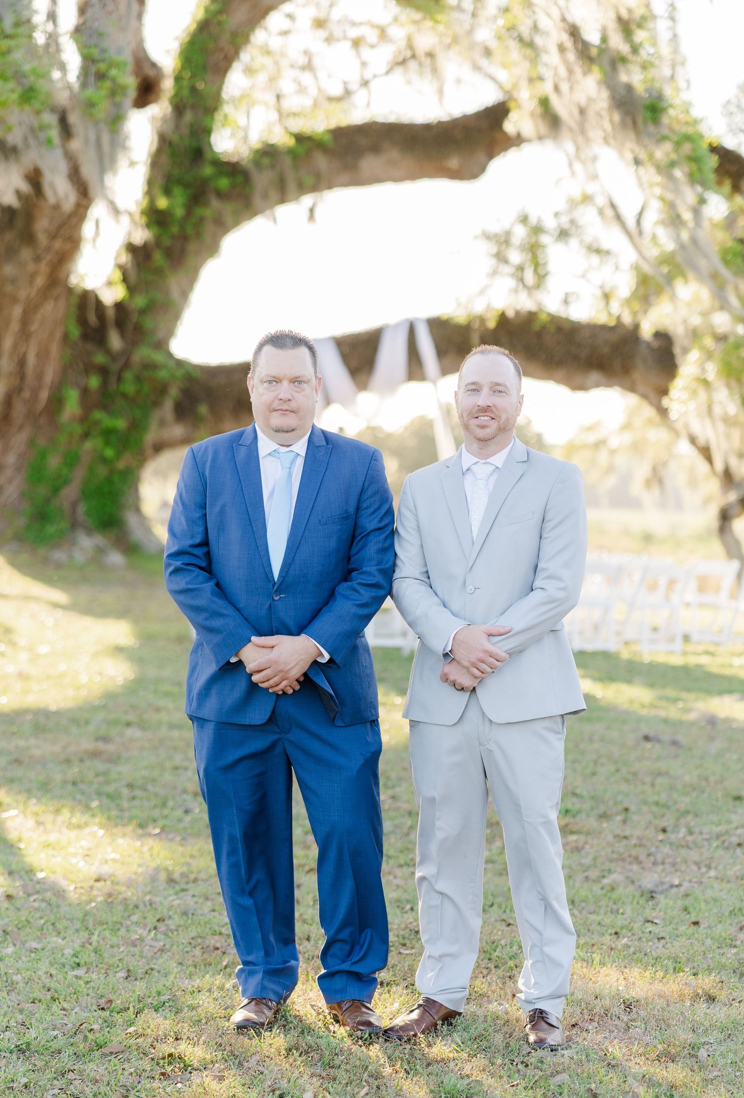 Two men in suits, one navy blue and one light gray, stand together at outdoor wedding ceremony venue.