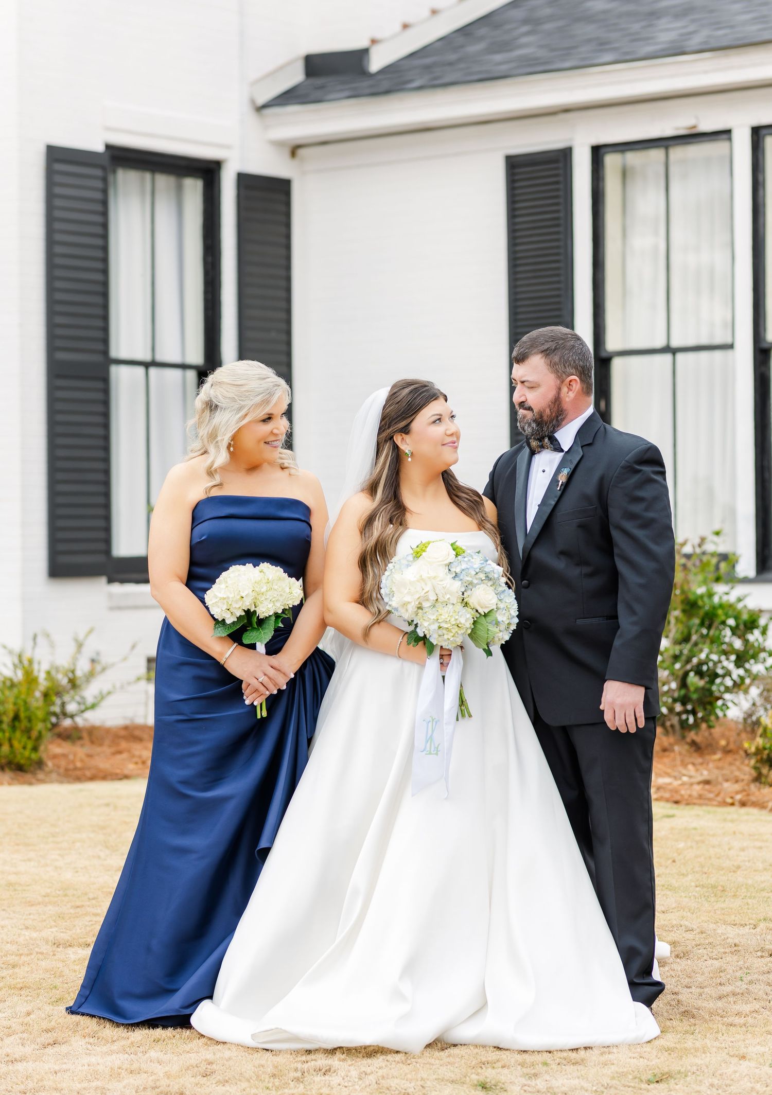 Bride in white ball gown holds white hydrangea bouquet flanked by parents in navy dress and black tuxedo.