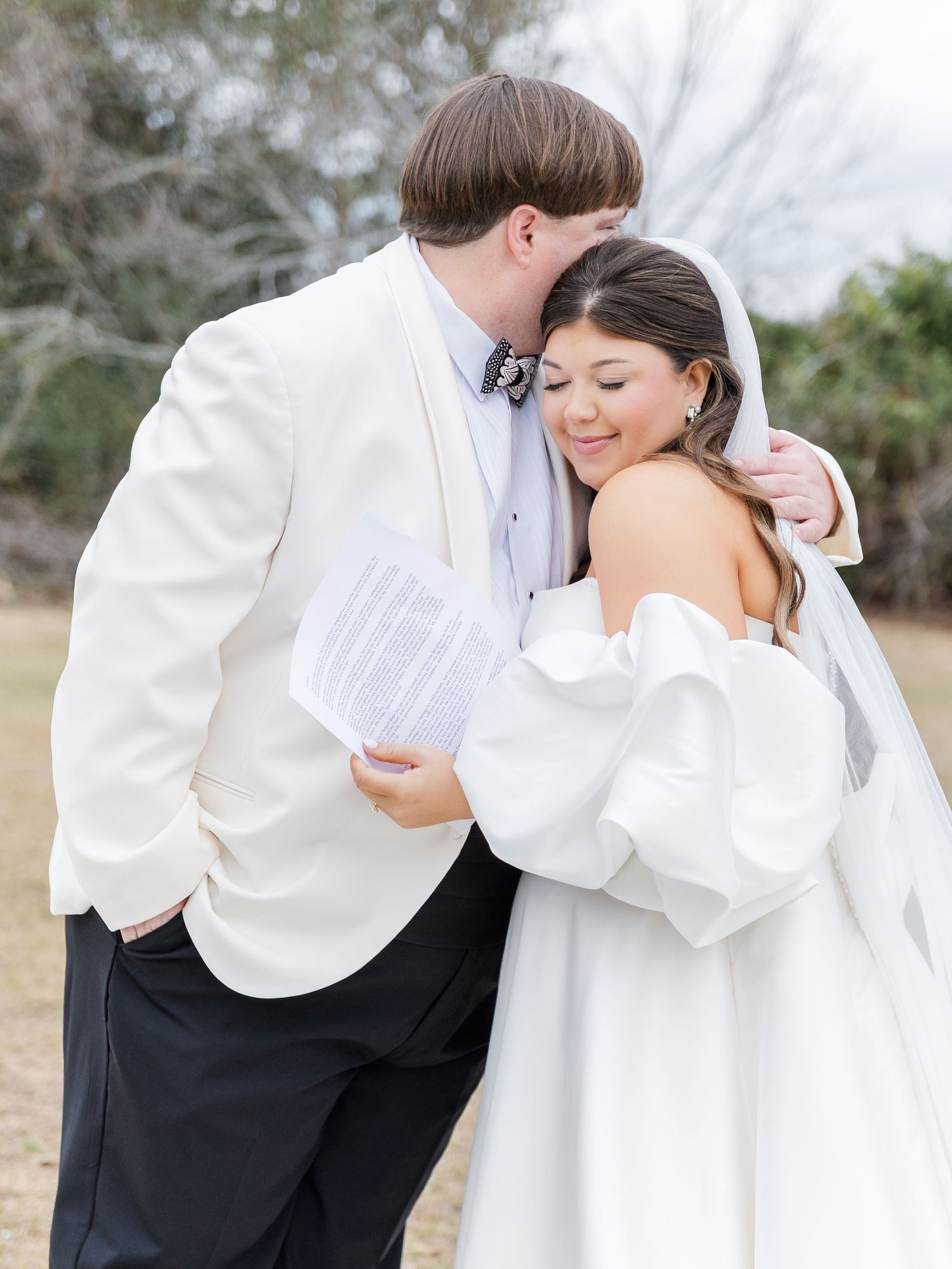 Groom in white tuxedo kisses bride's head while reading vows, bride smiles in off-shoulder wedding gown.