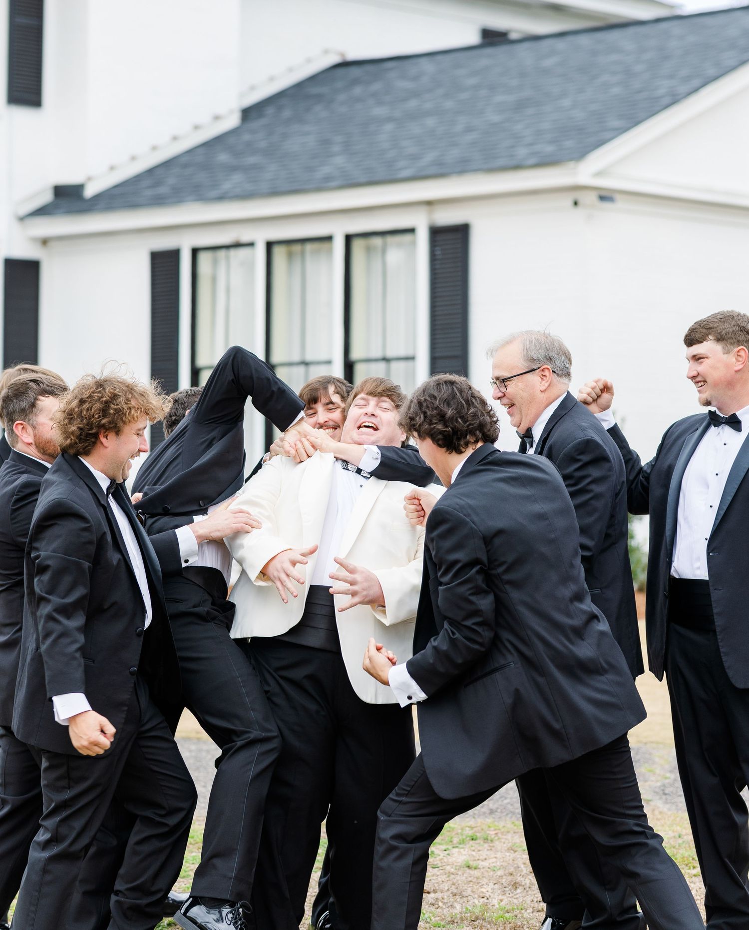 Groom in white jacket playfully lifted by groomsmen in black tuxedos outside white venue with black shutters.