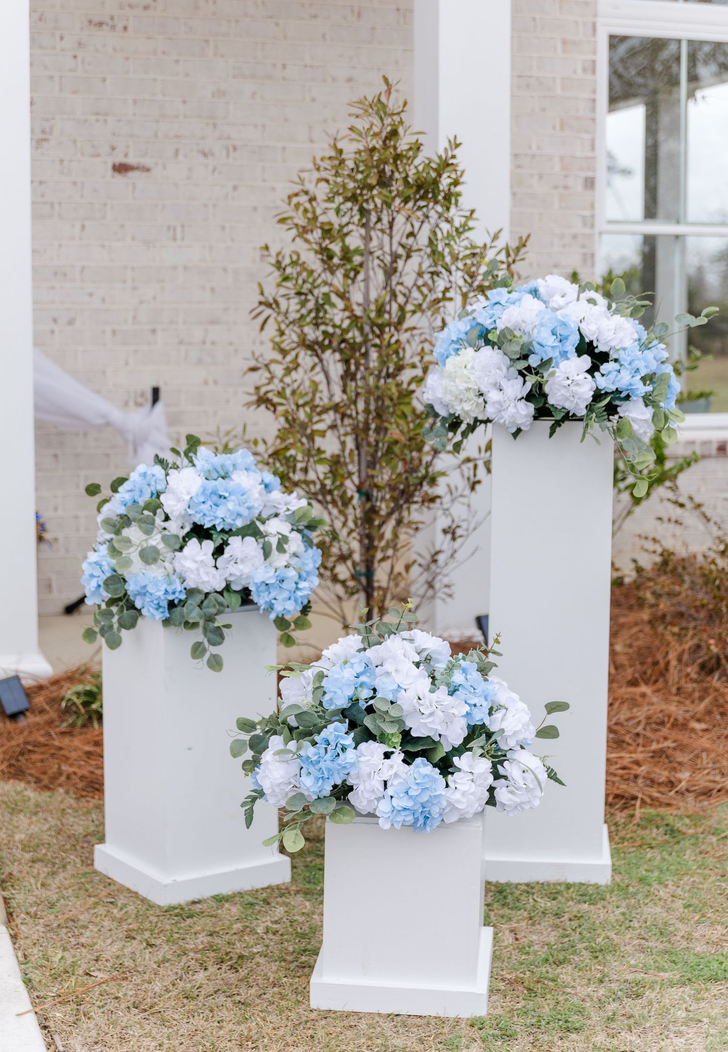 Three white pedestal stands adorned with blue and white hydrangea floral arrangements outside a brick wedding venue.