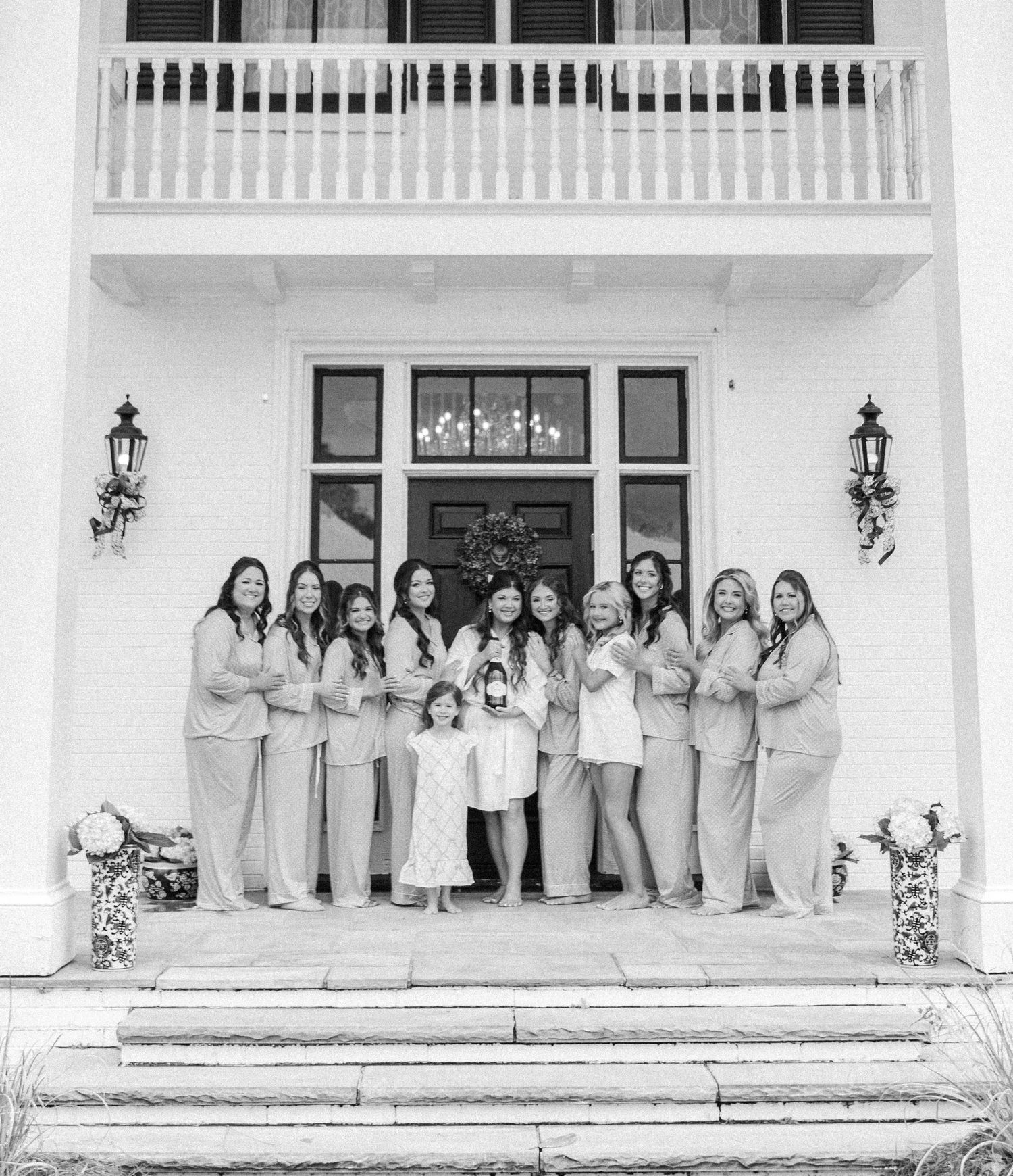 Black and white photo of bridal party in robes posing on mansion steps with flower girl holding bouquet.