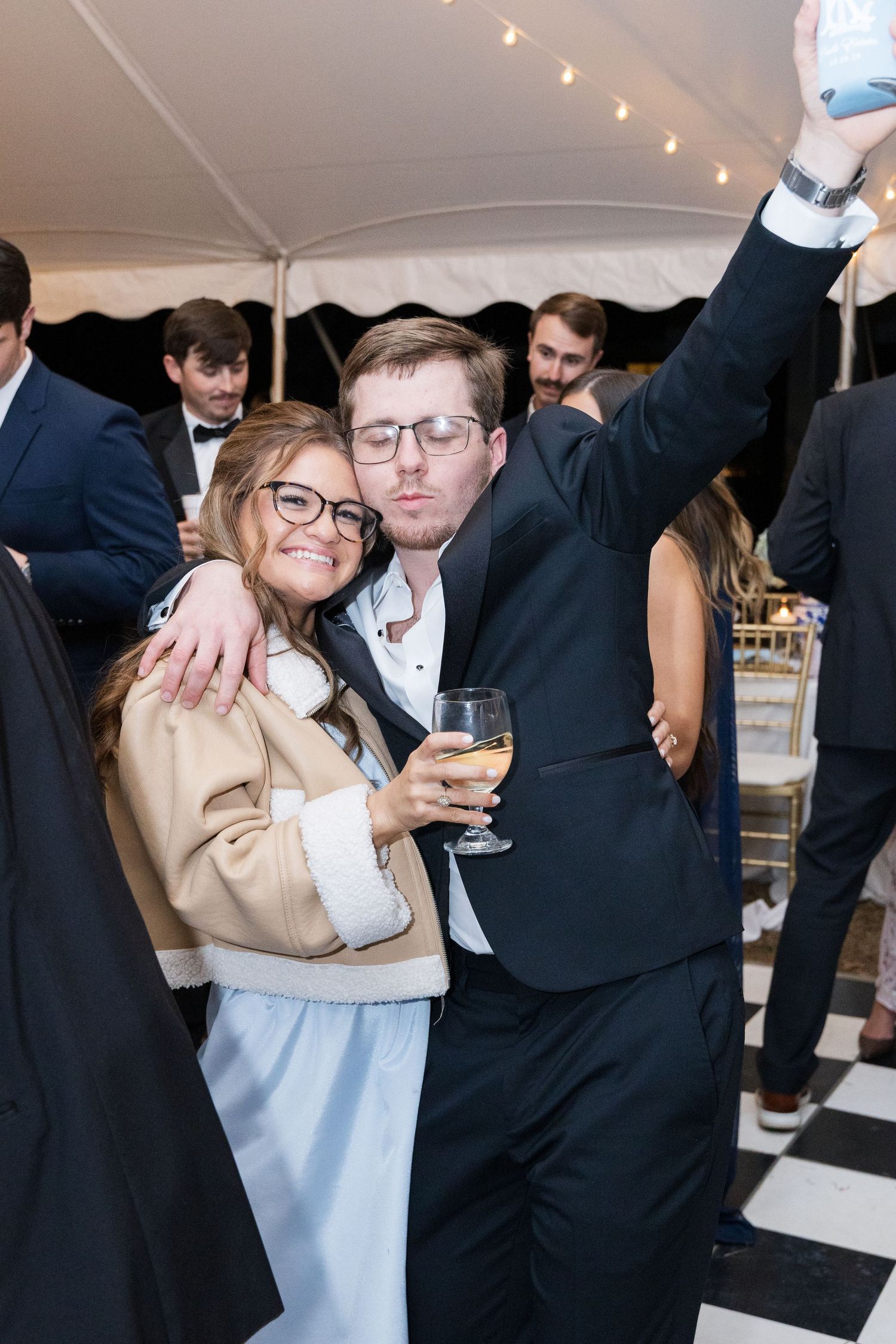 Couple in formal wear takes a selfie while holding wine glass at elegant tented wedding reception.