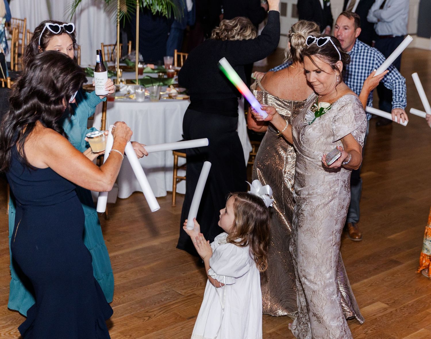 Flower girl in white dress dances with wedding guests holding colorful glow sticks at reception.