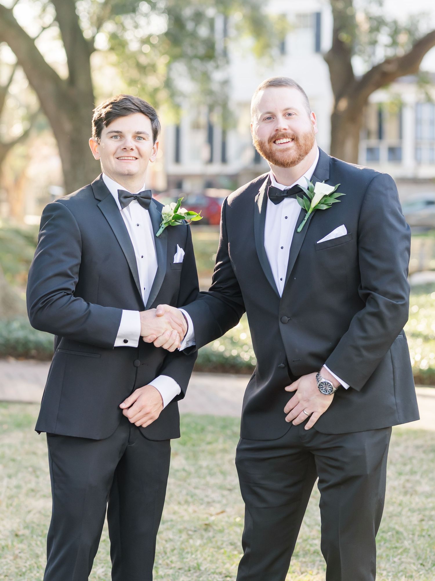 Two groomsmen in black tuxedos with bow ties and floral boutonnieres shaking hands outdoors.