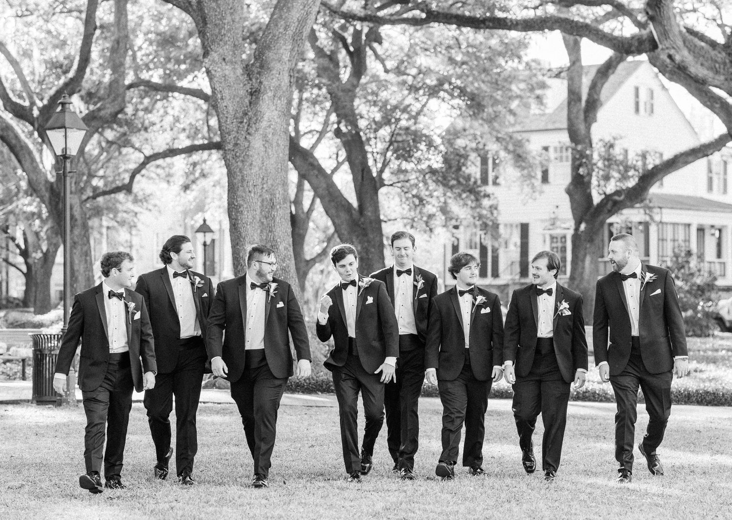 A group of men in black tuxedos walk together in a line on a tree-lined path in black and white photography.