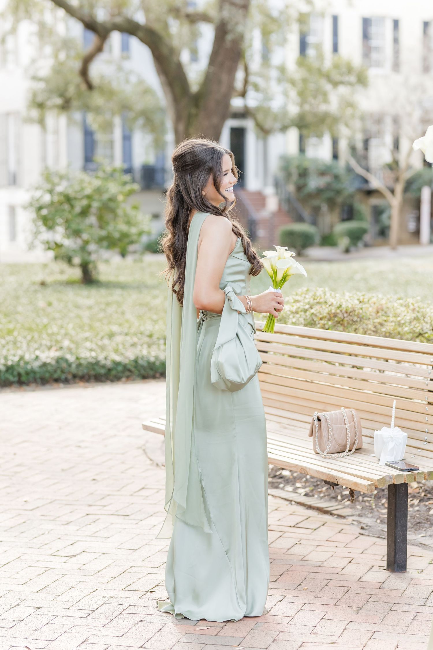 Person in sage green dress sits on wooden bench in sunlit courtyard.