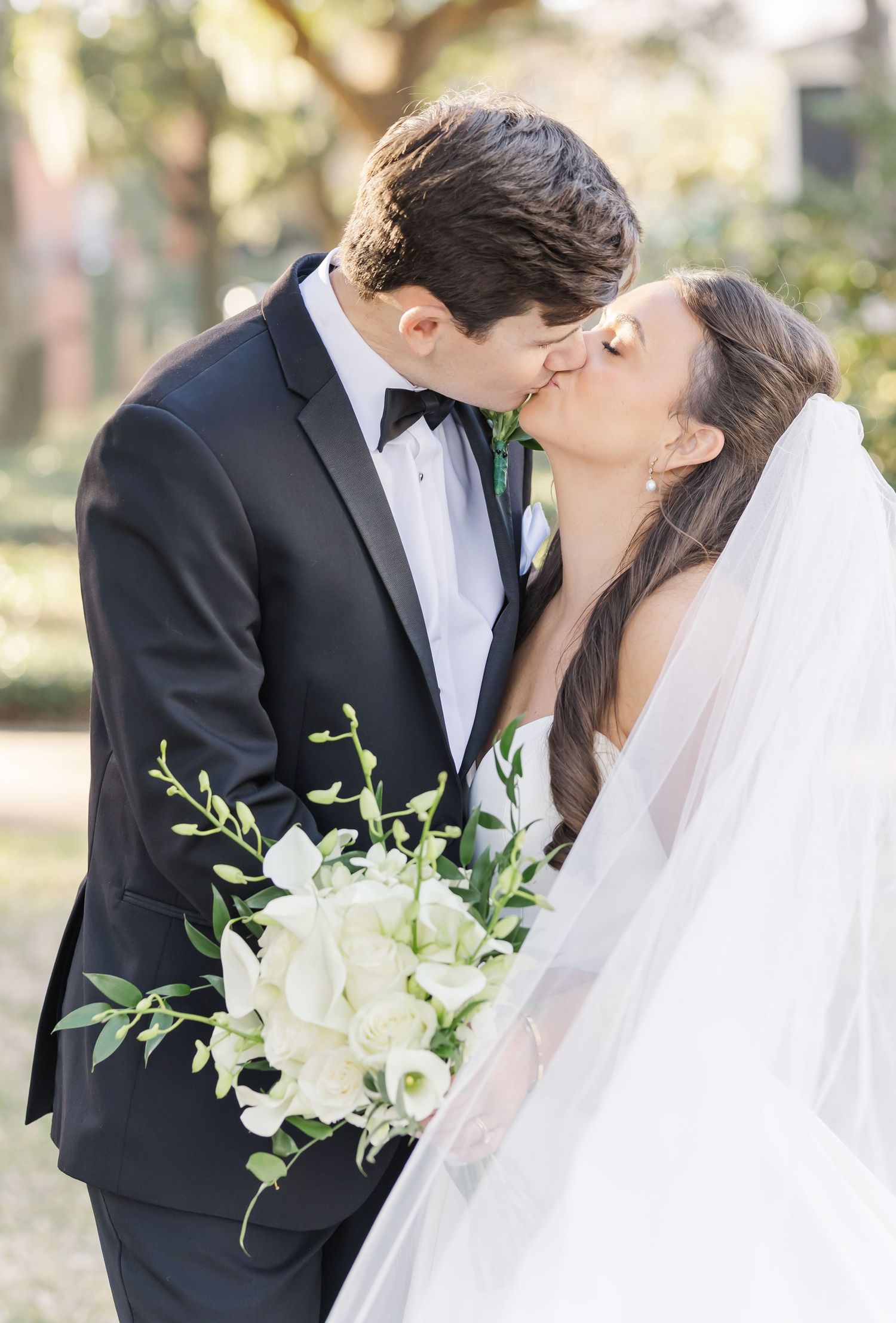 A romantic wedding moment outdoors with a white bouquet of flowers and flowing veil in natural lighting.