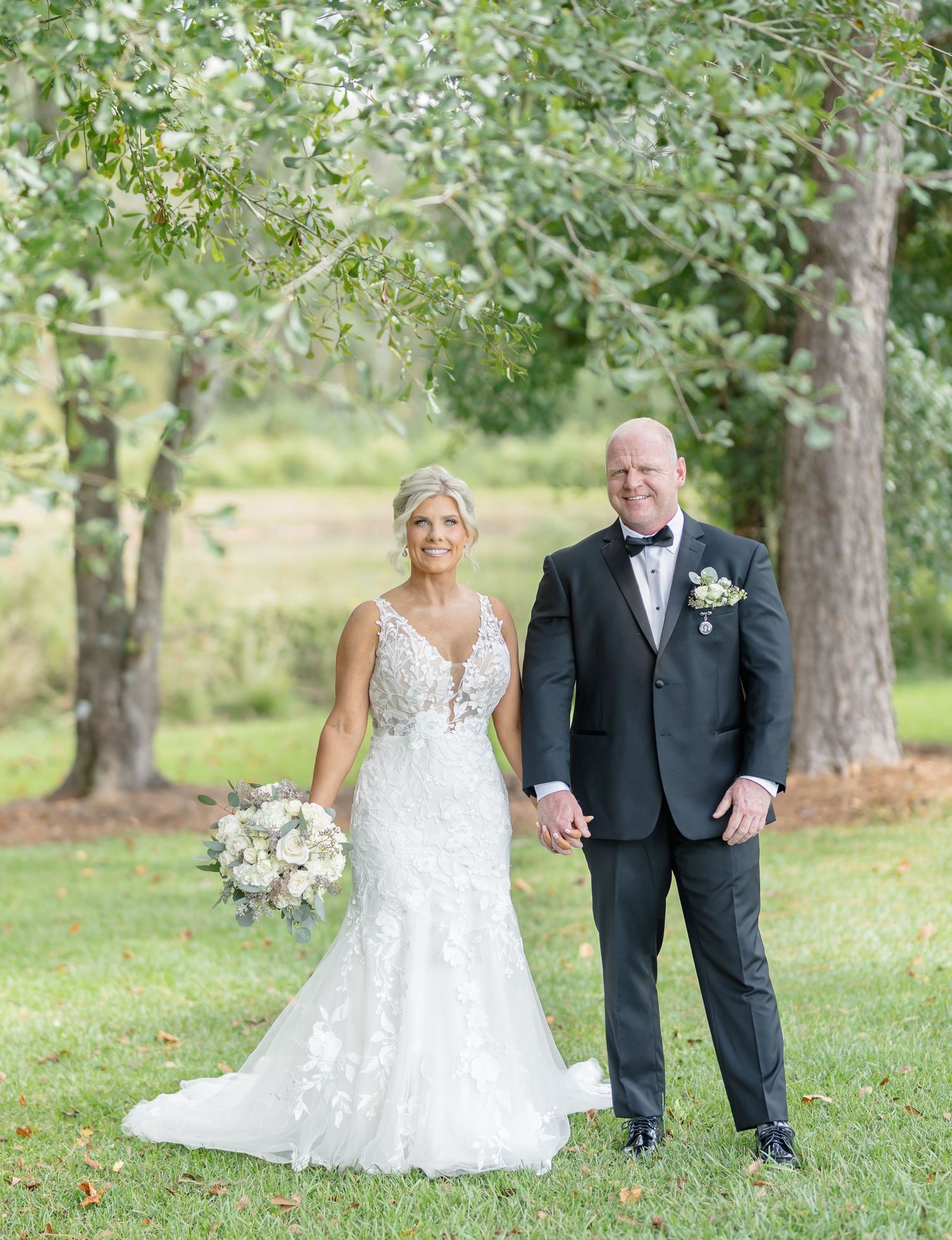 A couple in wedding attire walking together through a scenic outdoor garden setting with lush green trees.