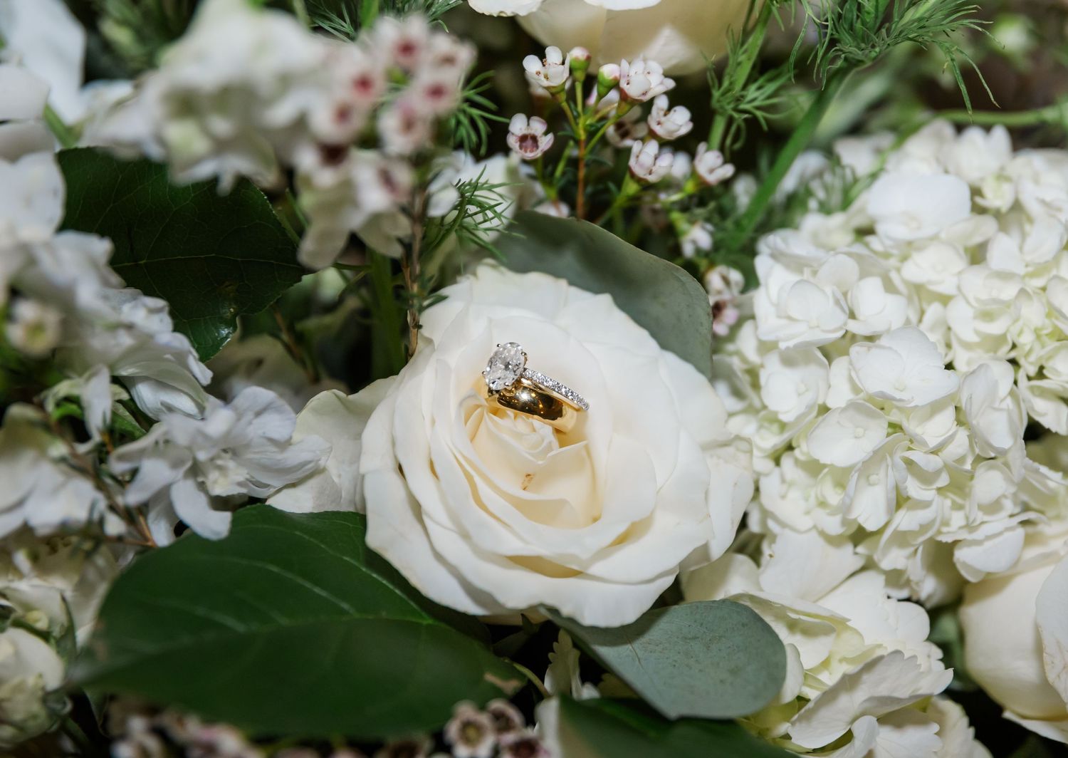 A delicate white rose and hydrangea bouquet with a gold wedding ring nestled in the center of the bloom.