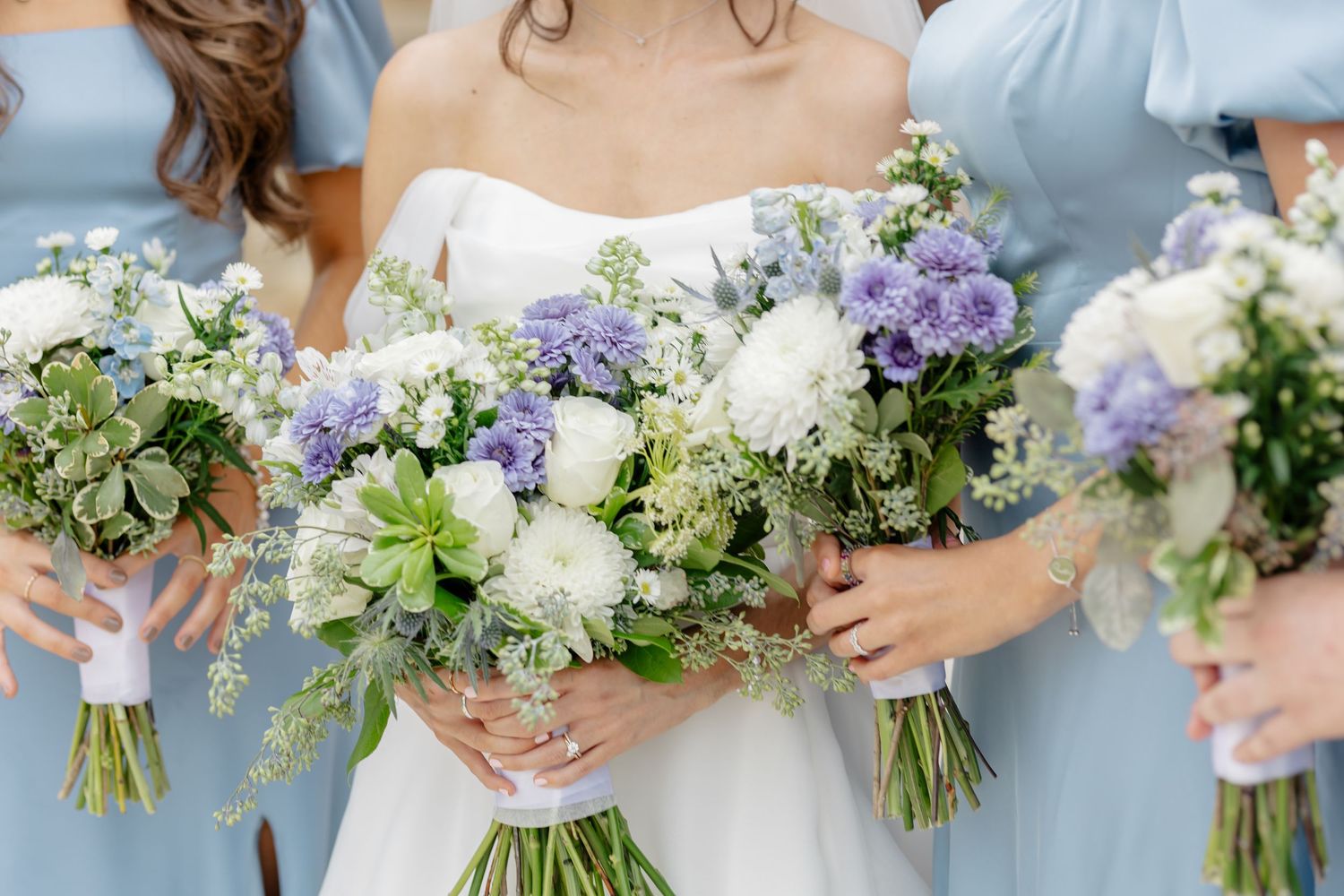 Elegant wedding bouquets featuring purple and white flowers with delicate greenery held by bridesmaids in blue dresses.