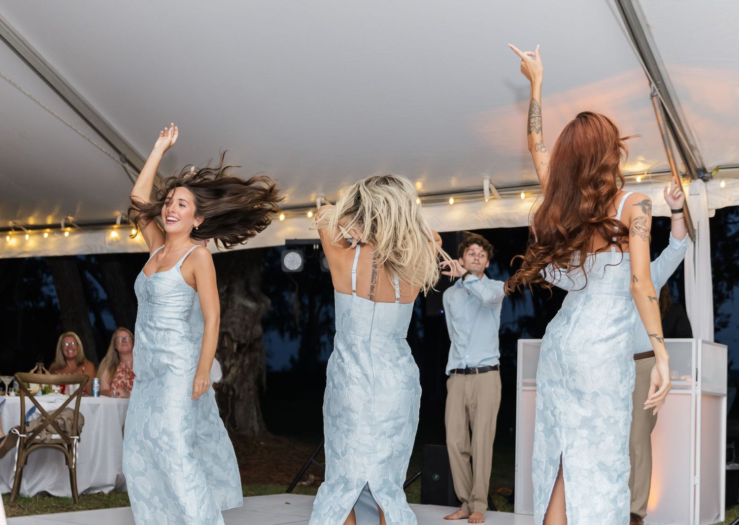 People dancing and celebrating with raised arms under a white tent at an evening event.