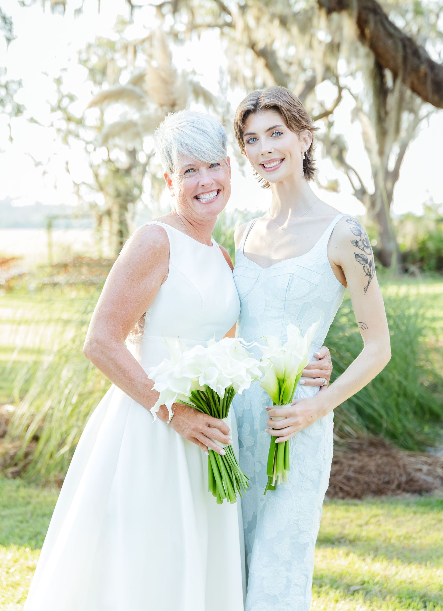 Two people in white and light blue dresses hold white calla lily bouquets while standing in a sunlit garden.