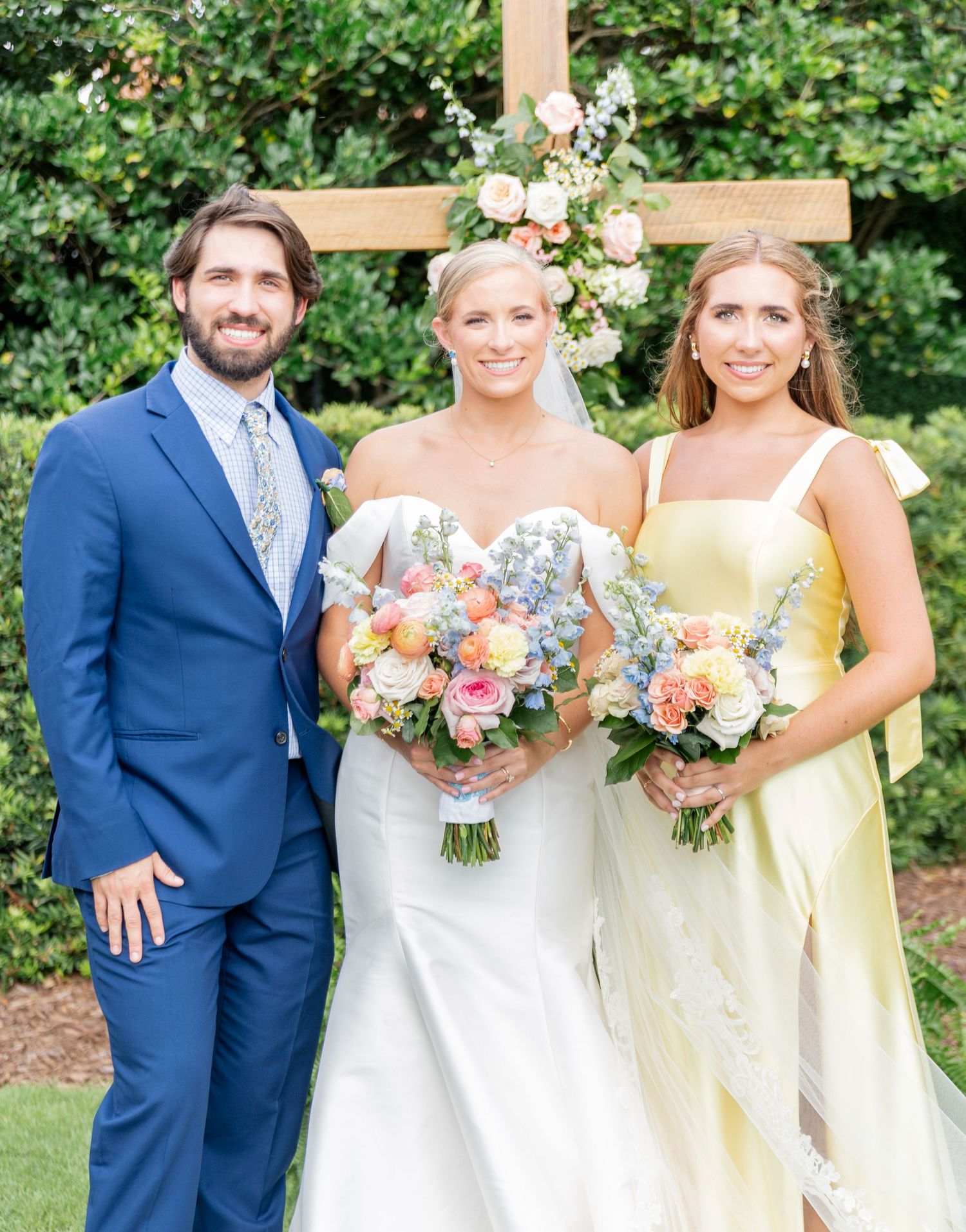 Wedding party poses outdoors with bridesmaids in pale yellow dresses and groomsmen in navy suits holding colorful bouquets.