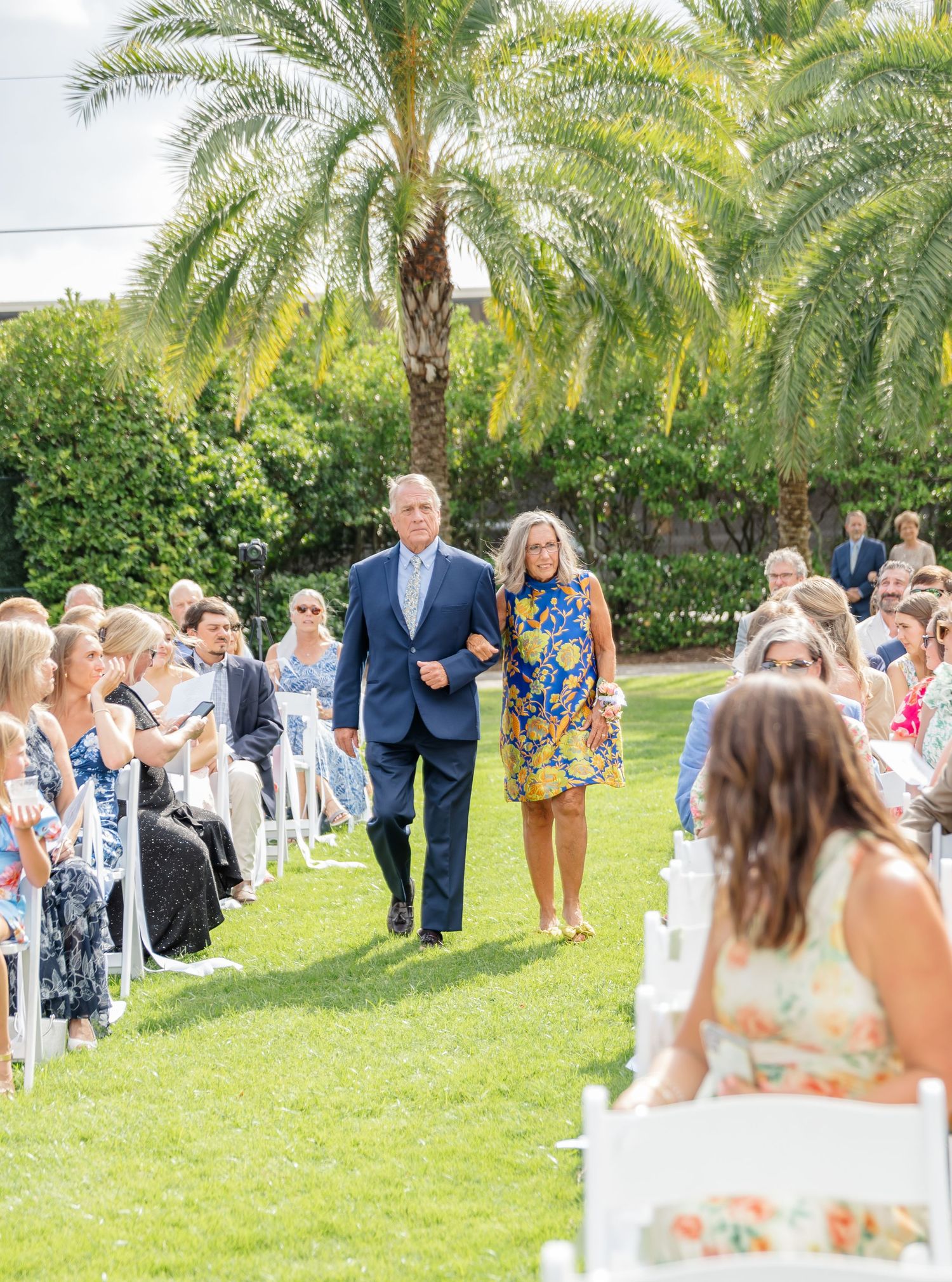 Outdoor wedding ceremony processional on green lawn with palm trees and guests seated in white chairs.