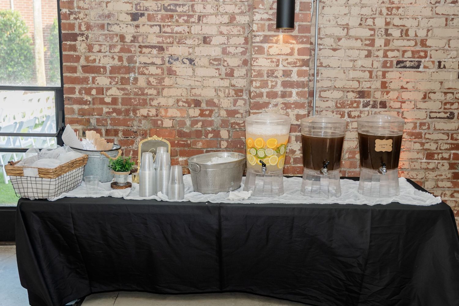 A beverage station with glass dispensers and a black tablecloth set against an exposed brick wall.