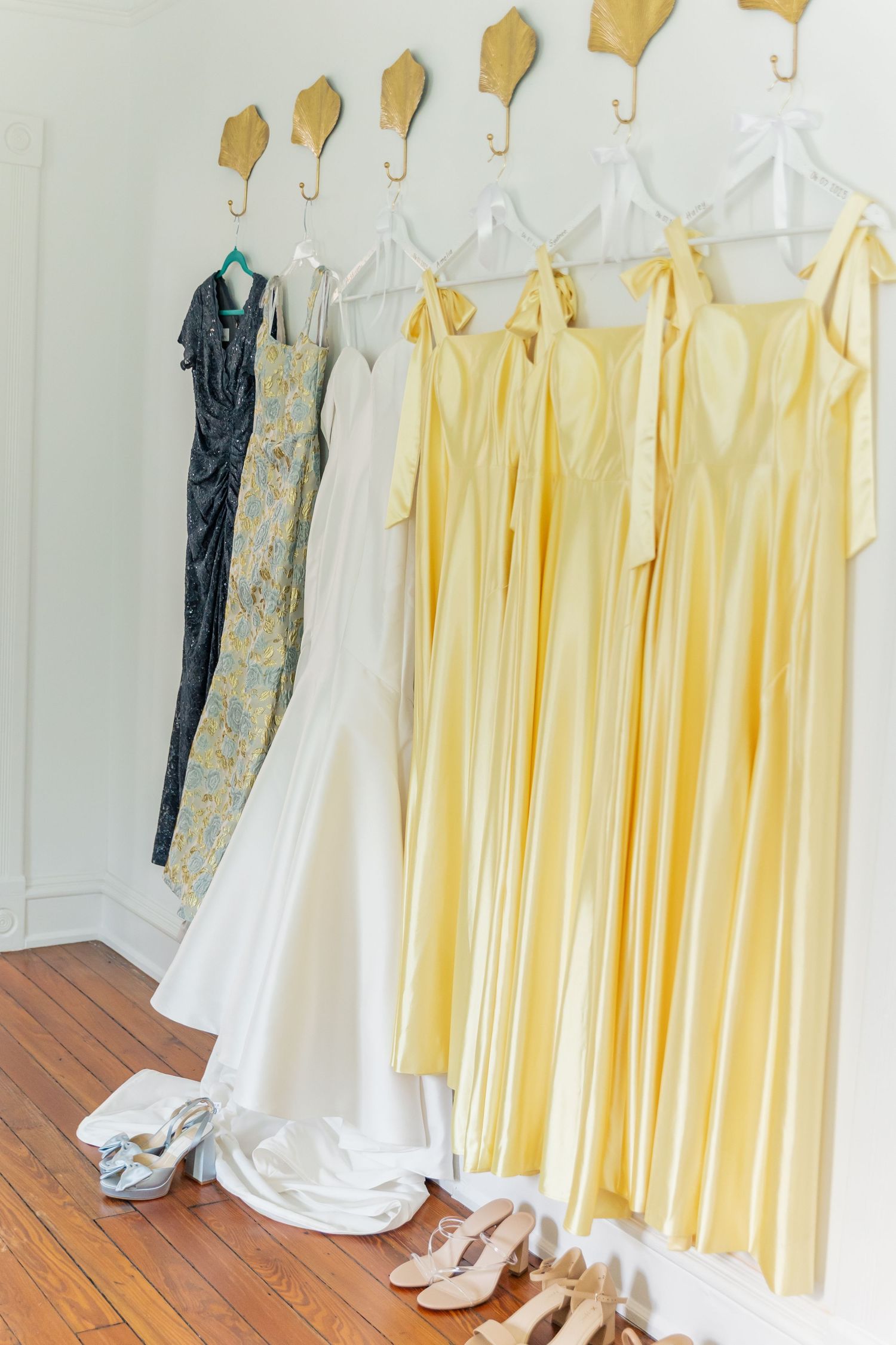 Wedding party dresses in yellow and white arranged in a row with a navy suit and formal shoes on wooden floor.