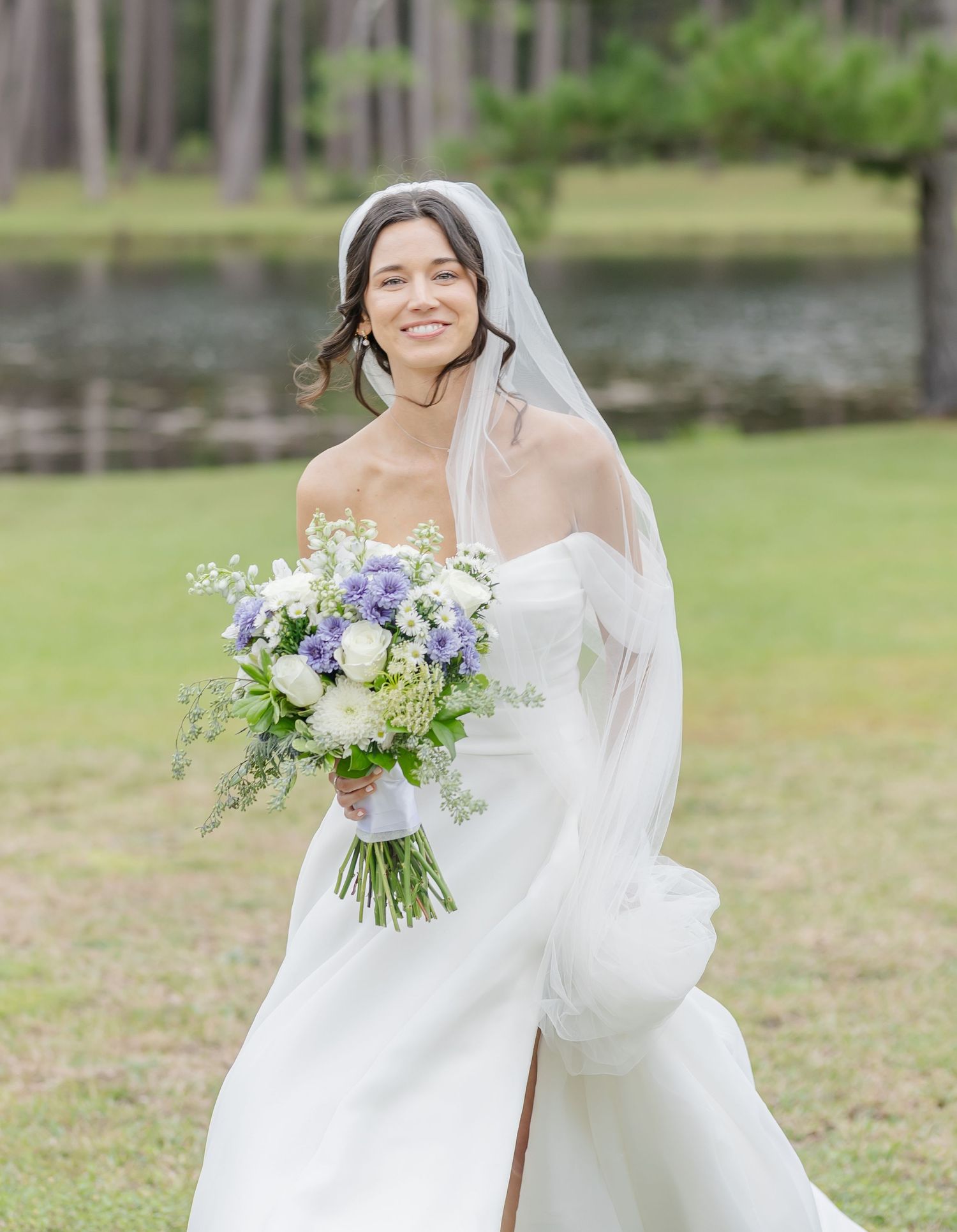 Bride in flowing white gown holds delicate bouquet of purple and white flowers in an outdoor garden setting.
