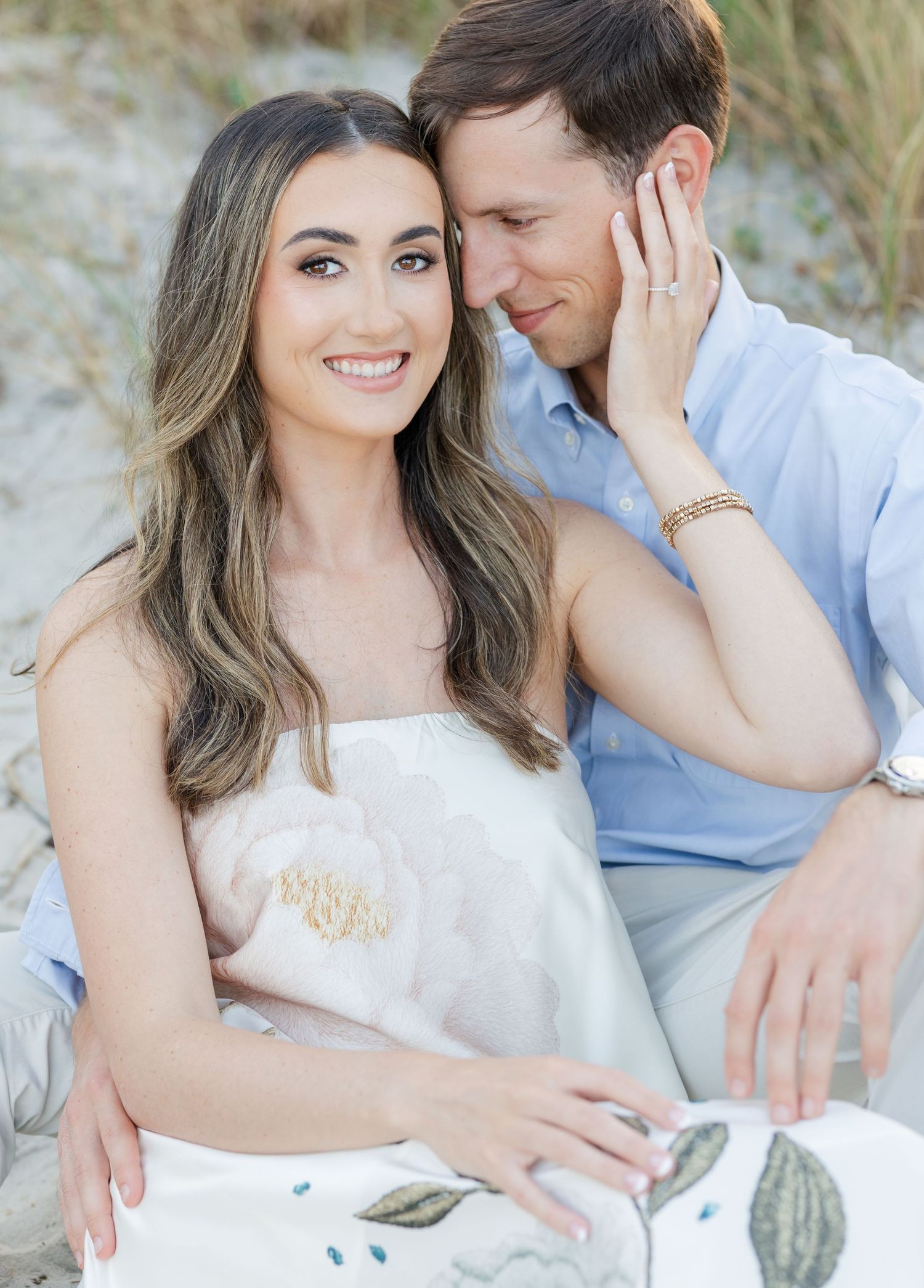 Couple sitting on beach sand dunes, woman in floral strapless dress smiling, man in blue shirt nuzzling her cheek.