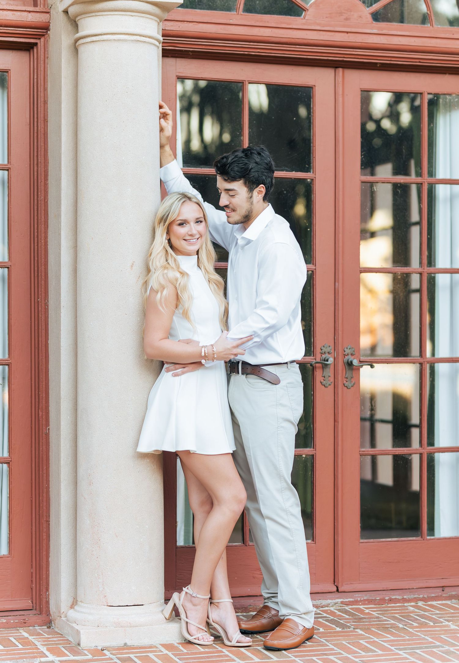 Couple posing by a column in front of red French doors, woman in white mini dress, man in light gray trousers.