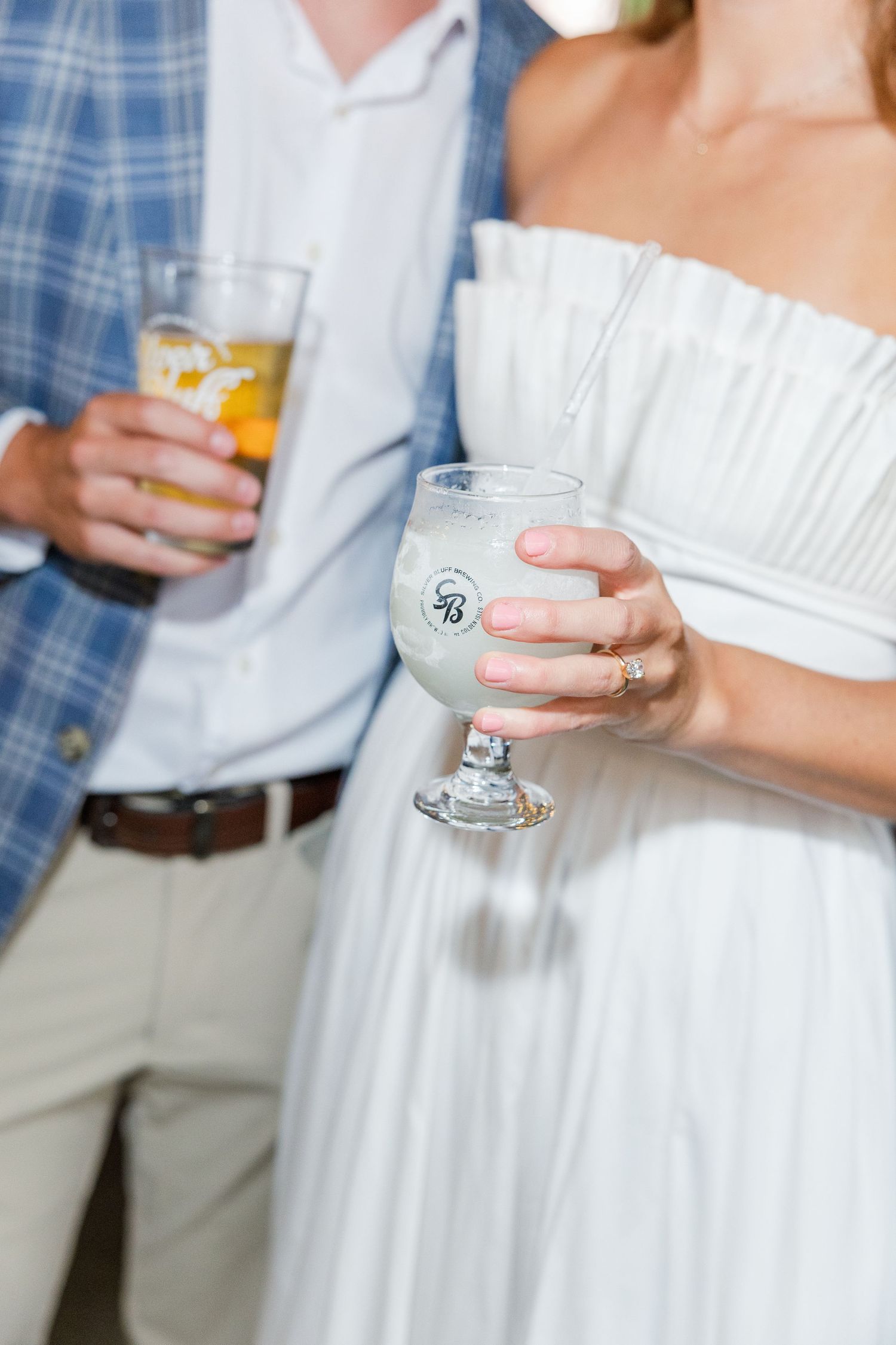 Couple holding drinks at a wedding, bride in white strapless dress with a branded goblet glass.
