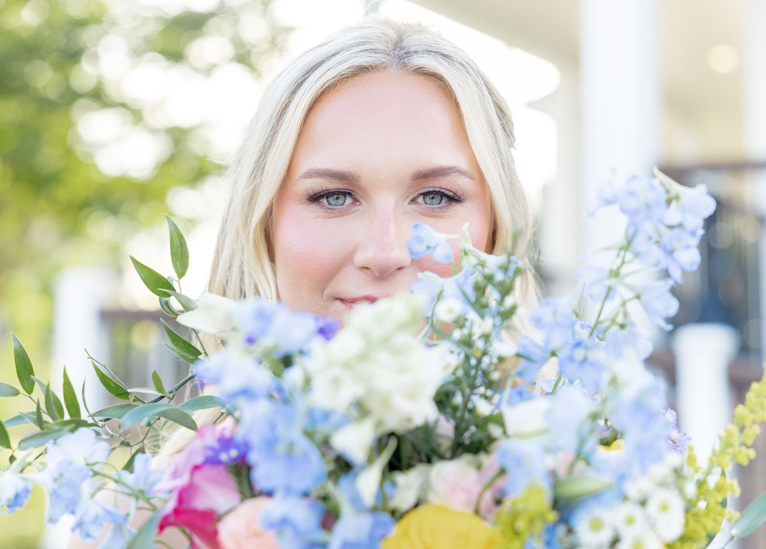 Beautiful blonde bride peeks over vibrant blue and pink wedding bouquet with sparkling blue eyes.
