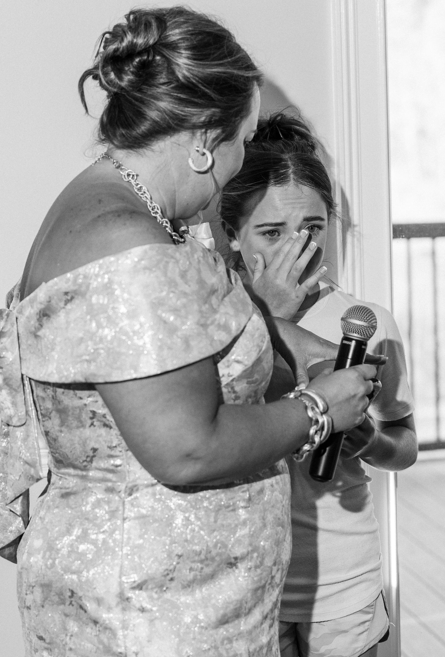 Black and white photo of woman comforting emotional crying bride holding microphone indoors.