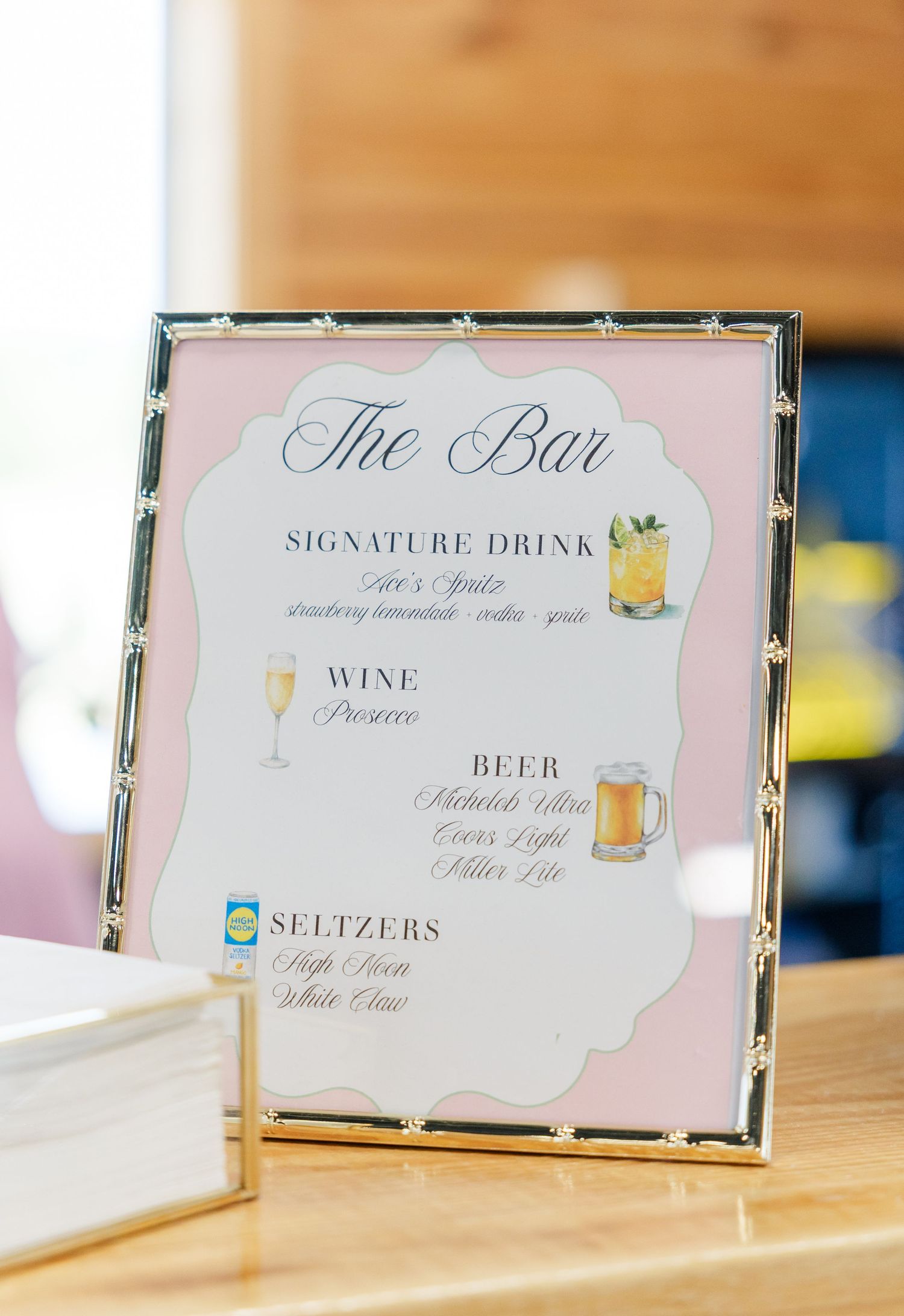 Framed wedding bar menu sign listing signature drinks, wine, beer, and seltzers in pink and white design.