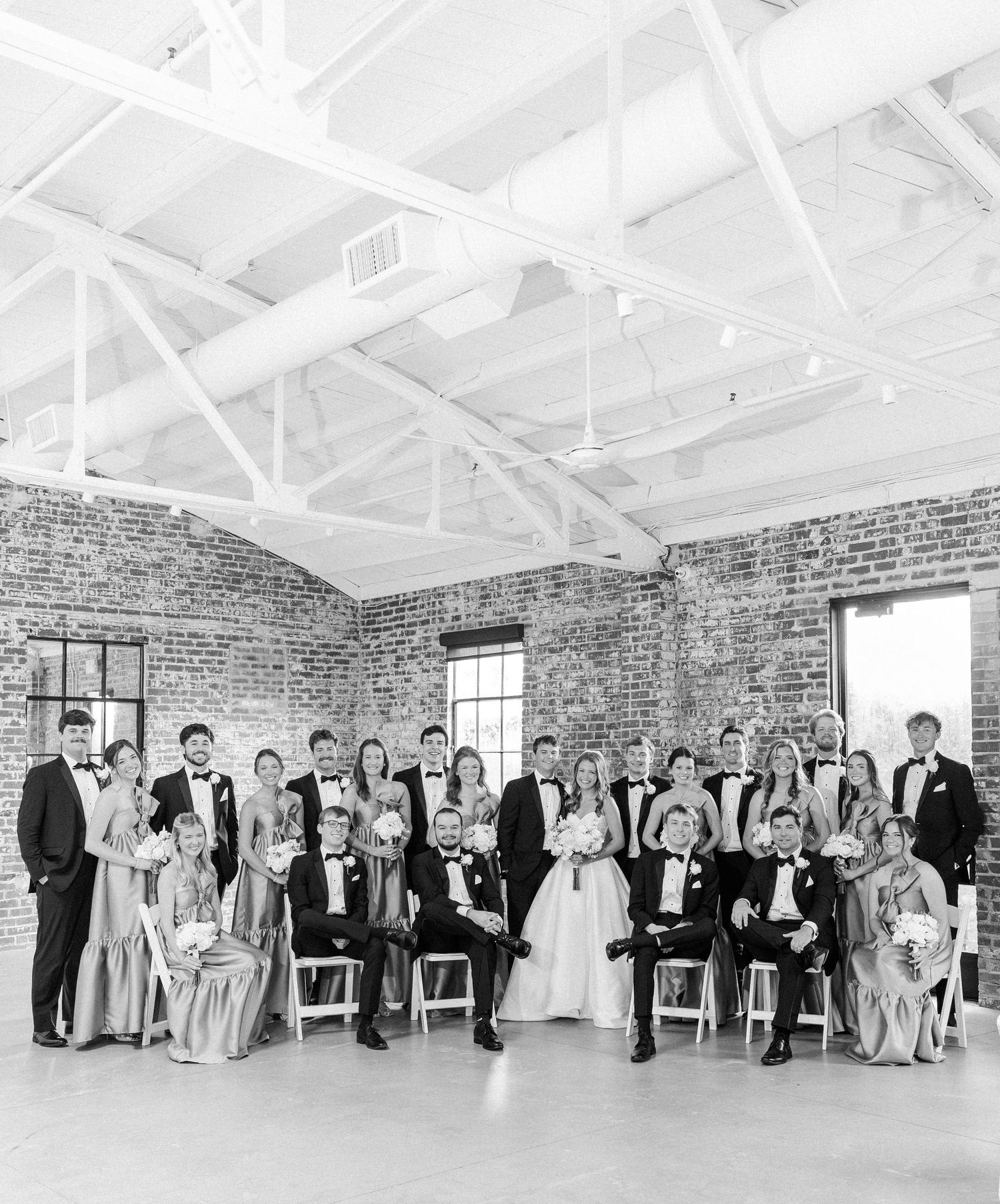 Large wedding party poses together in a rustic brick-walled venue with white exposed beam ceilings.