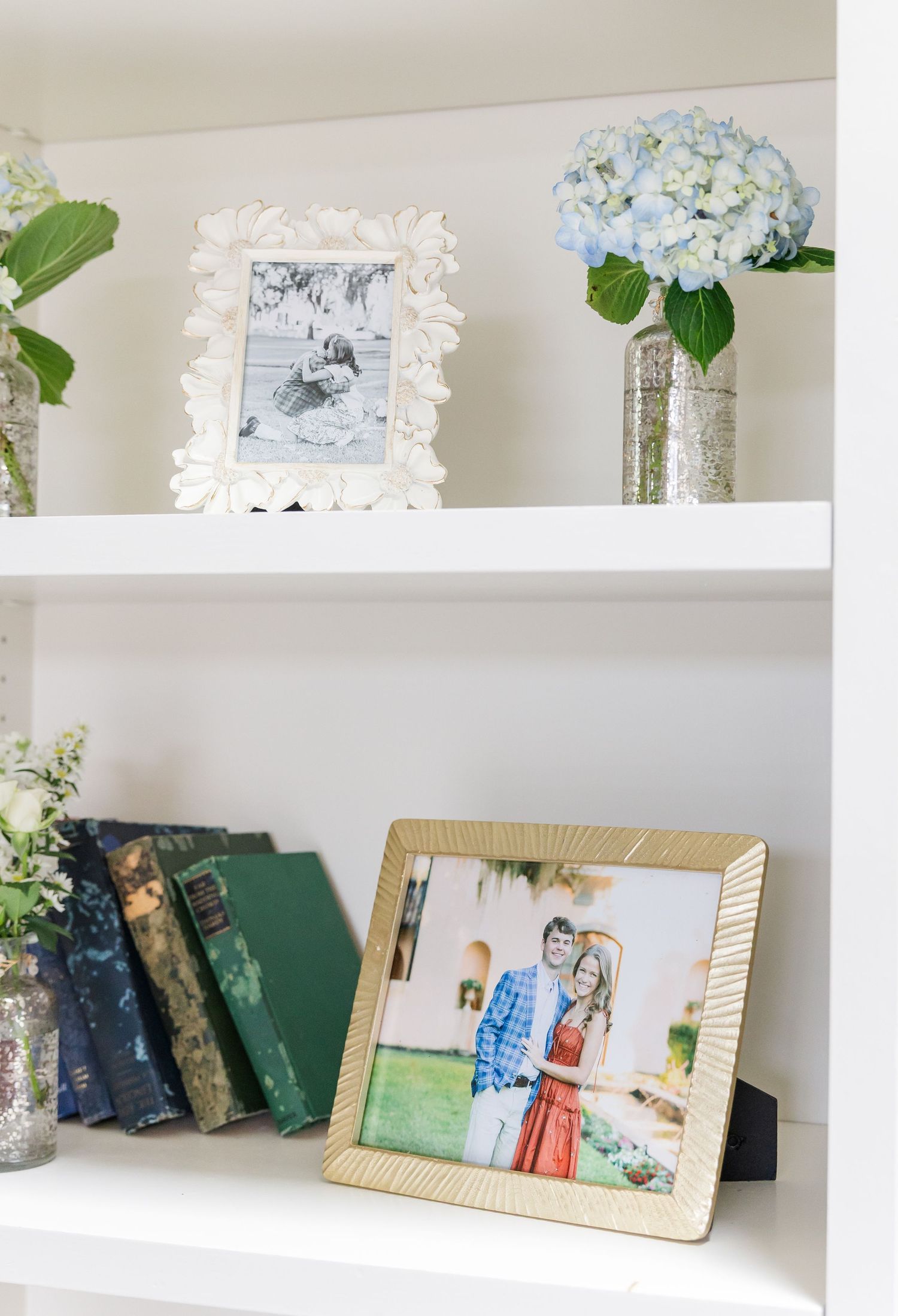 White built-in shelves styled with framed photos, vintage books, blue hydrangeas in vases, and a gold-framed couples portrait.