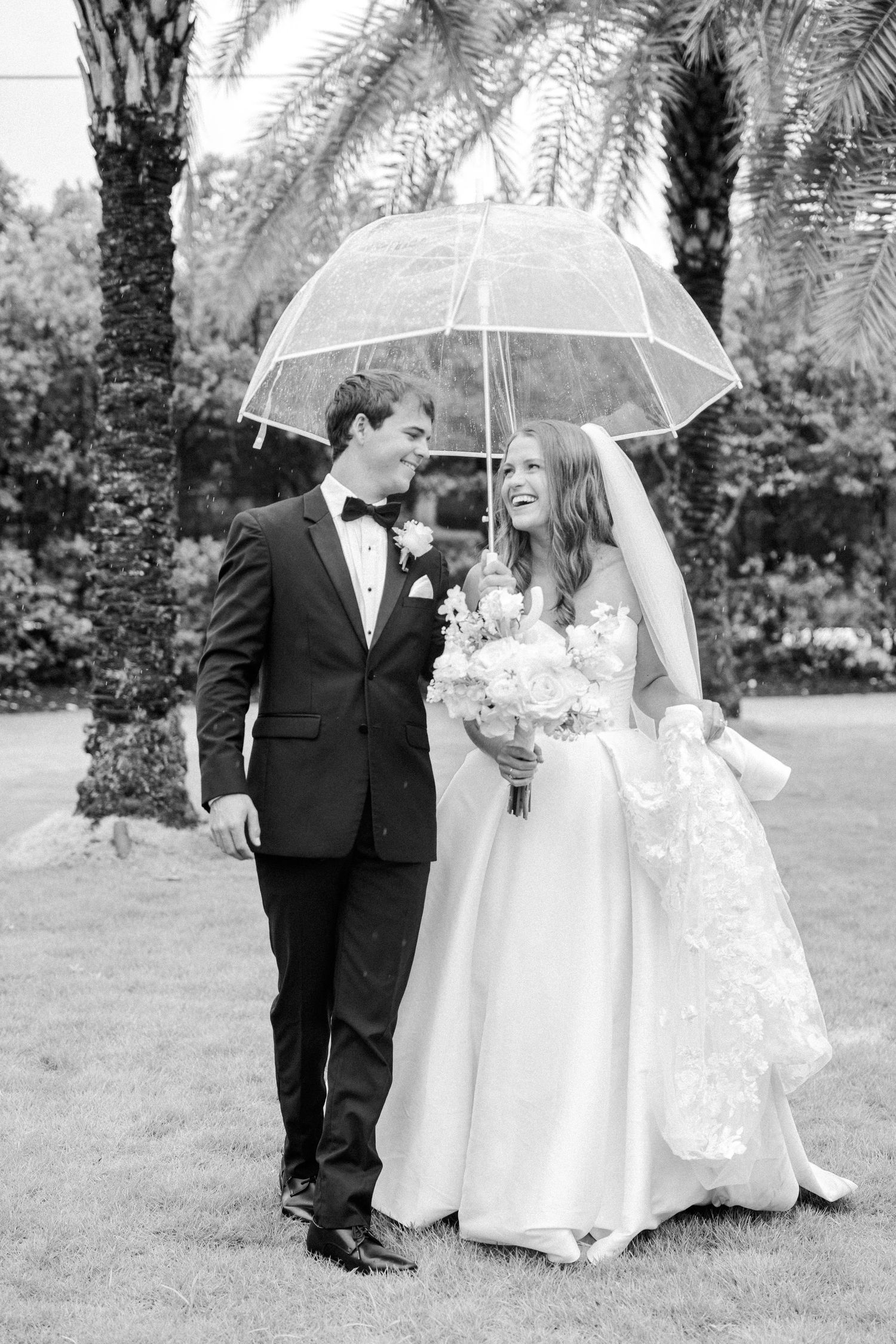 Black and white photo of bride and groom walking under umbrella among palm trees.