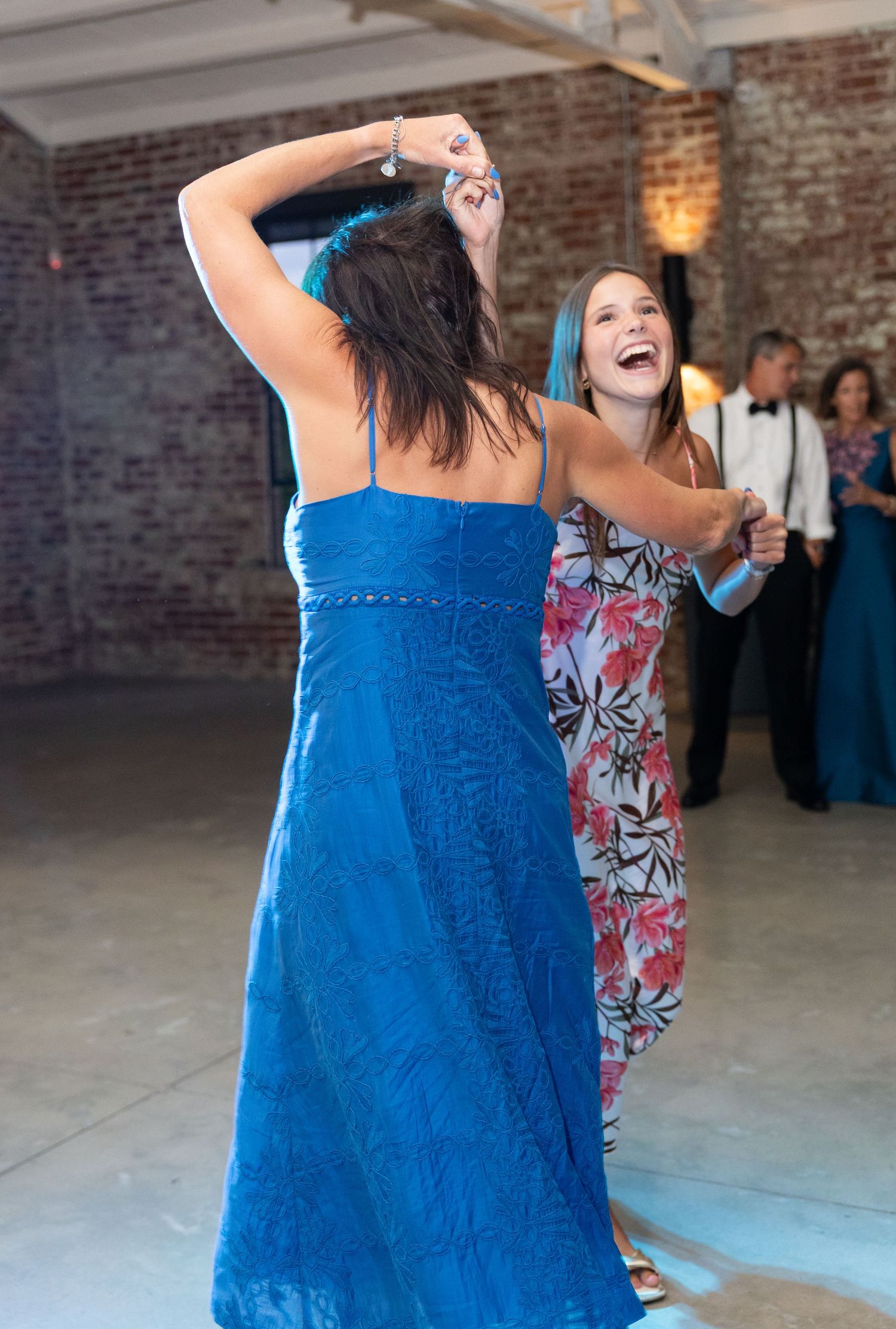 Two women in blue dresses laugh and dance together joyfully at wedding reception.