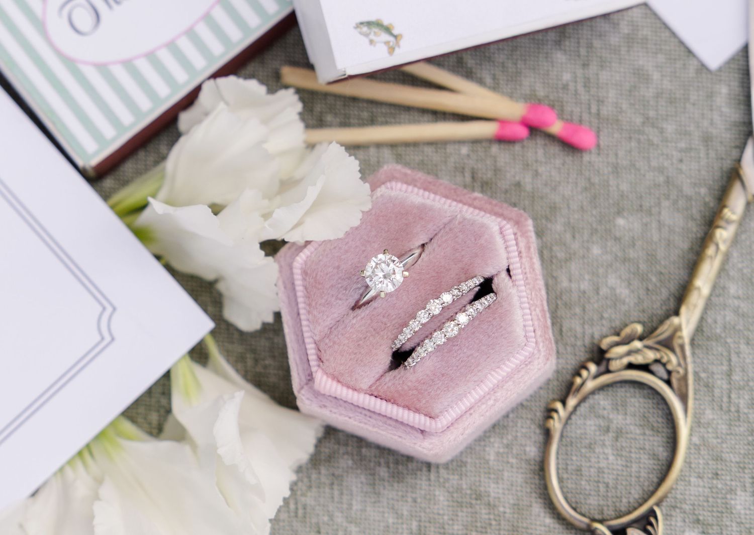 Two diamond rings displayed in a pink velvet hexagon ring box beside white flowers and gold scissors.