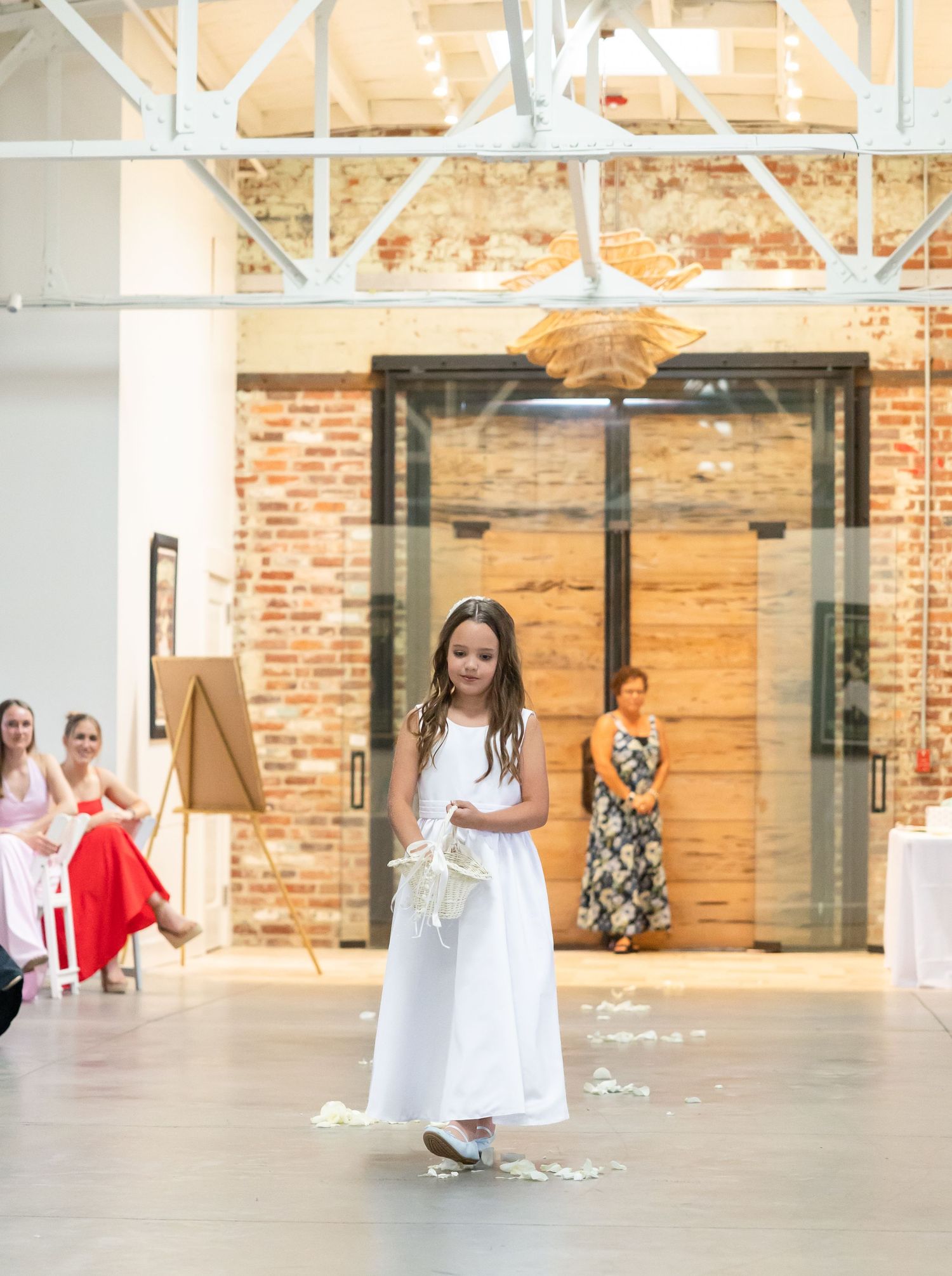 Young flower girl in white dress scatters petals down the aisle in a bright industrial wedding venue.