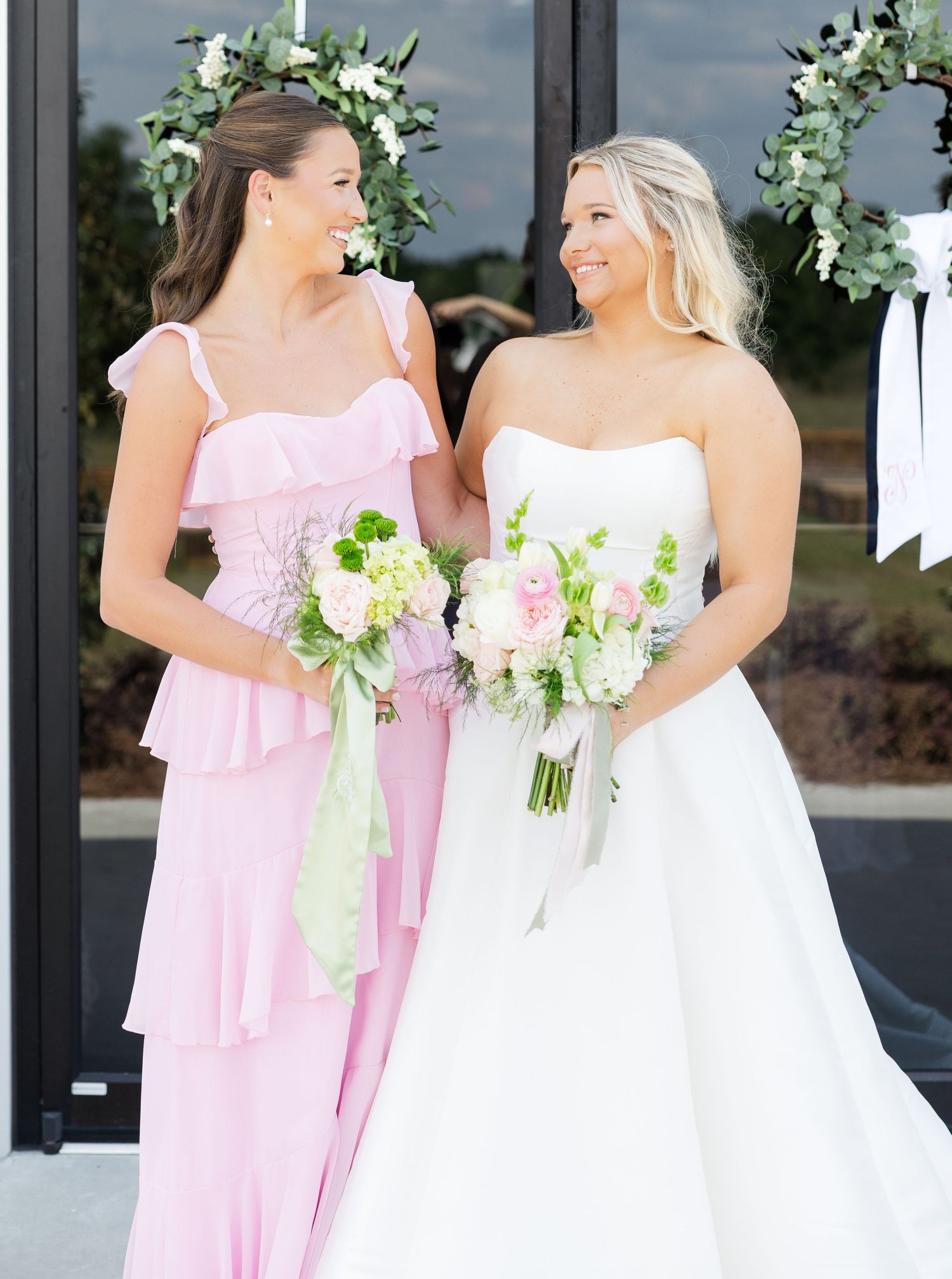 Bride in white strapless gown and bridesmaid in pink ruffle dress holding floral bouquets outdoors.
