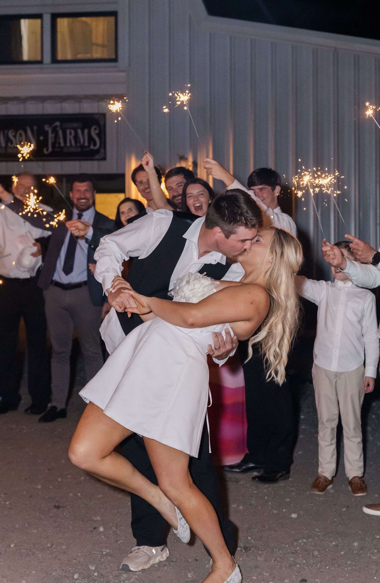 Bride and groom share a romantic dip kiss surrounded by guests holding sparklers at outdoor wedding reception.