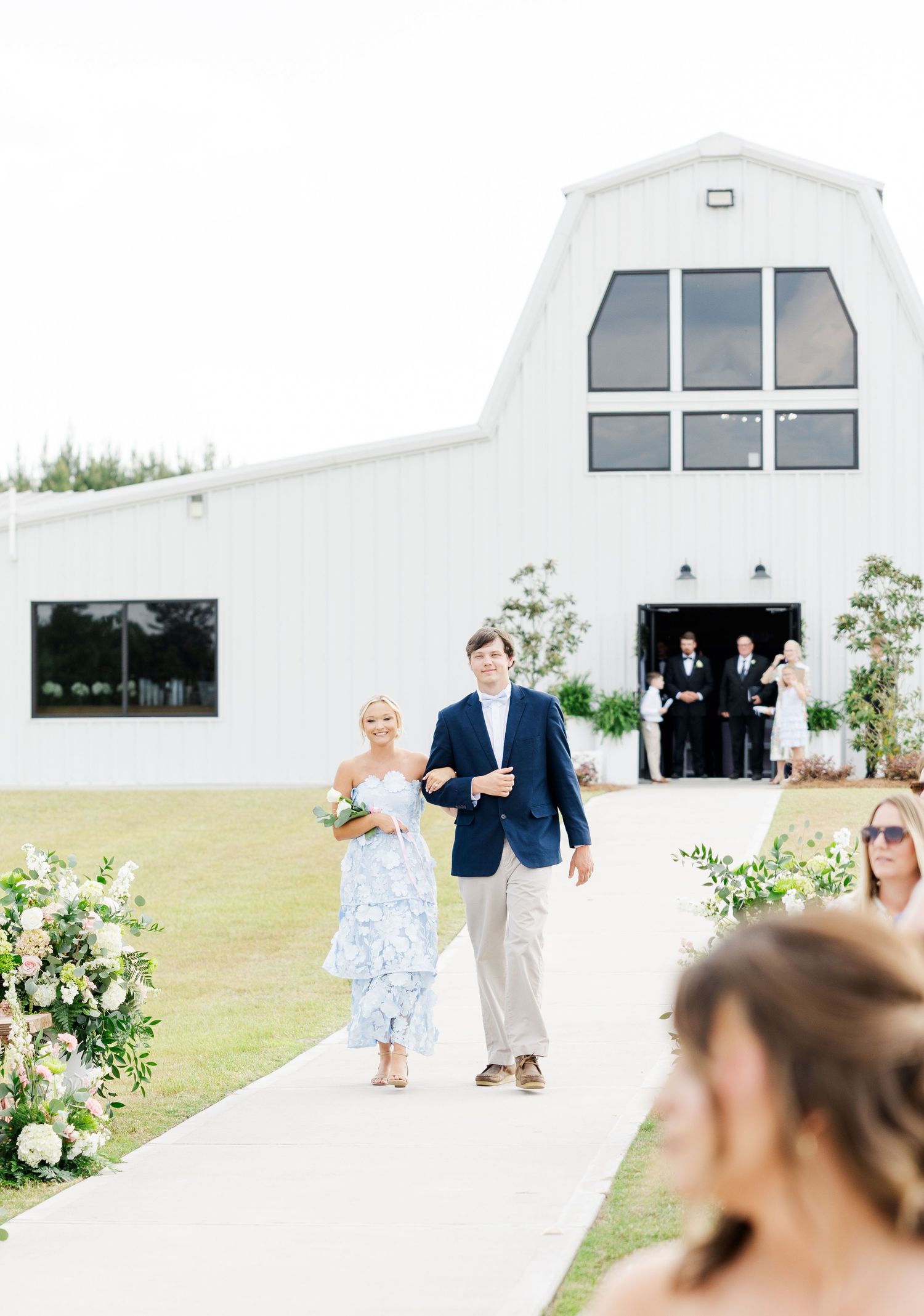 Couple walks down outdoor aisle toward white barn wedding venue surrounded by floral arrangements.