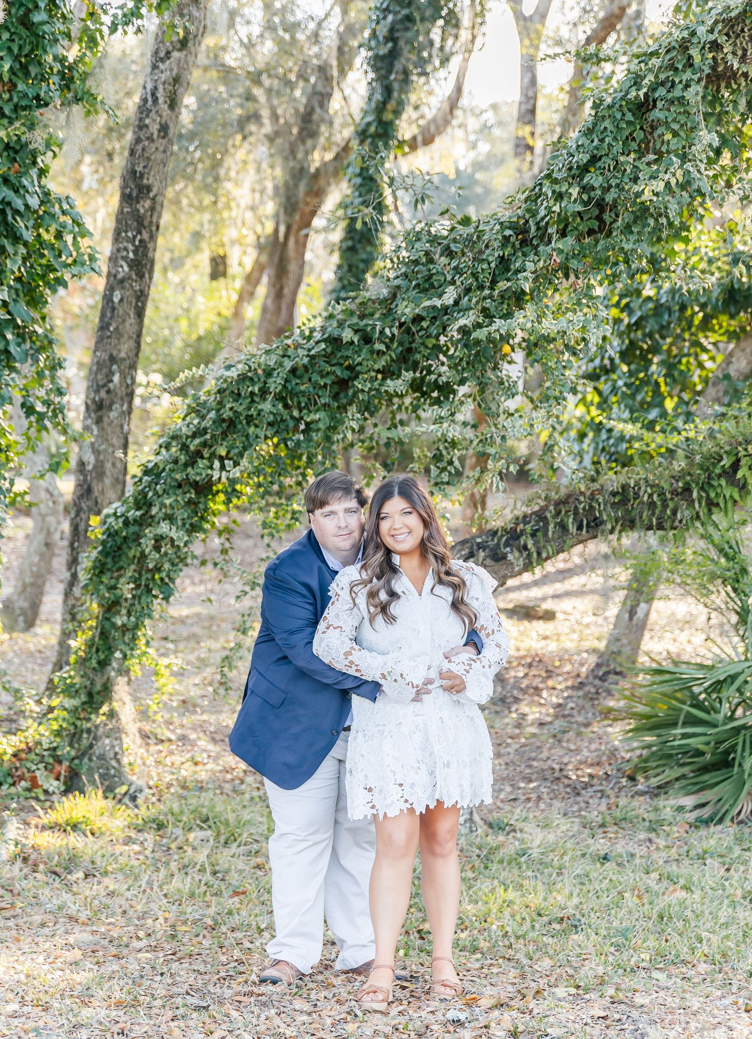 A couple embraces on a tree-lined path, wearing coordinated formal attire.