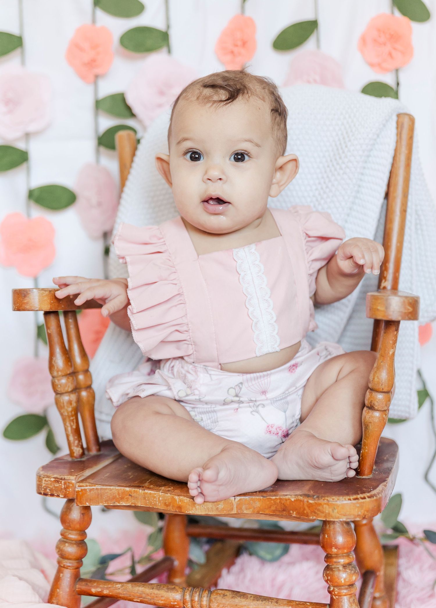 Smiling baby in pink outfit poses on vintage wooden chair with floral background.