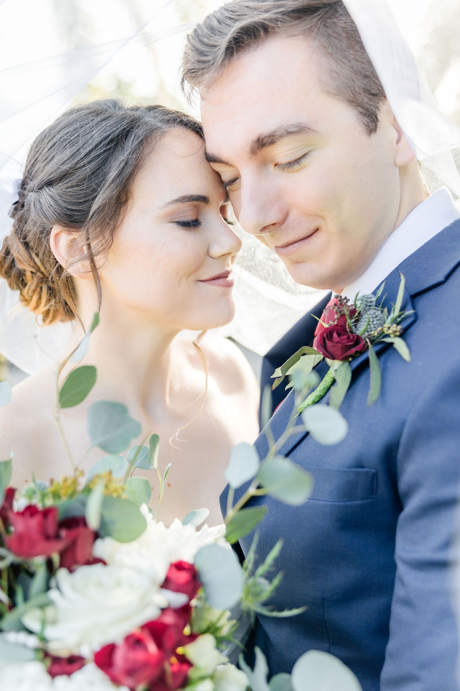 Intimate wedding portrait with red roses and eucalyptus bouquet in soft lighting.