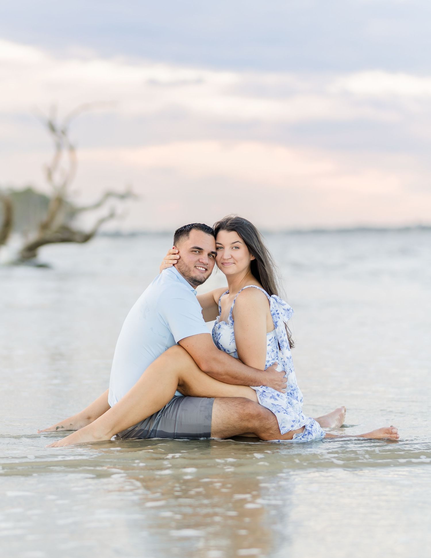 A couple sits together in shallow ocean water during sunset, creating a romantic beach photoshoot moment.
