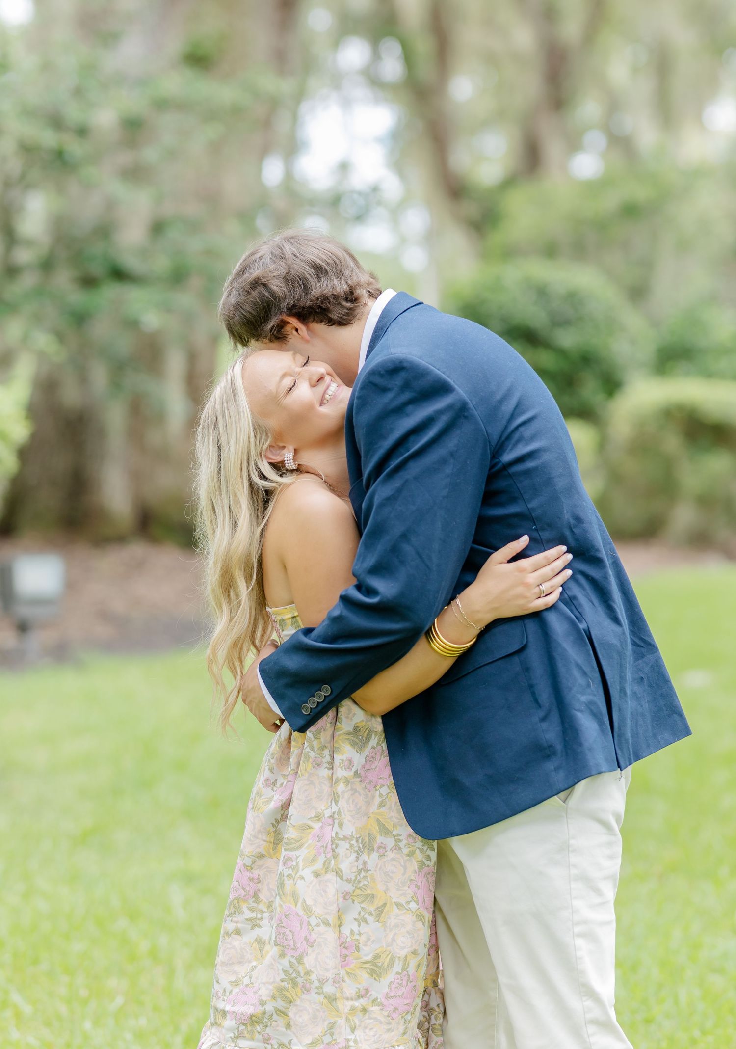 A romantic couple shares a tender embrace in a park setting with Spanish moss draped trees in the background.